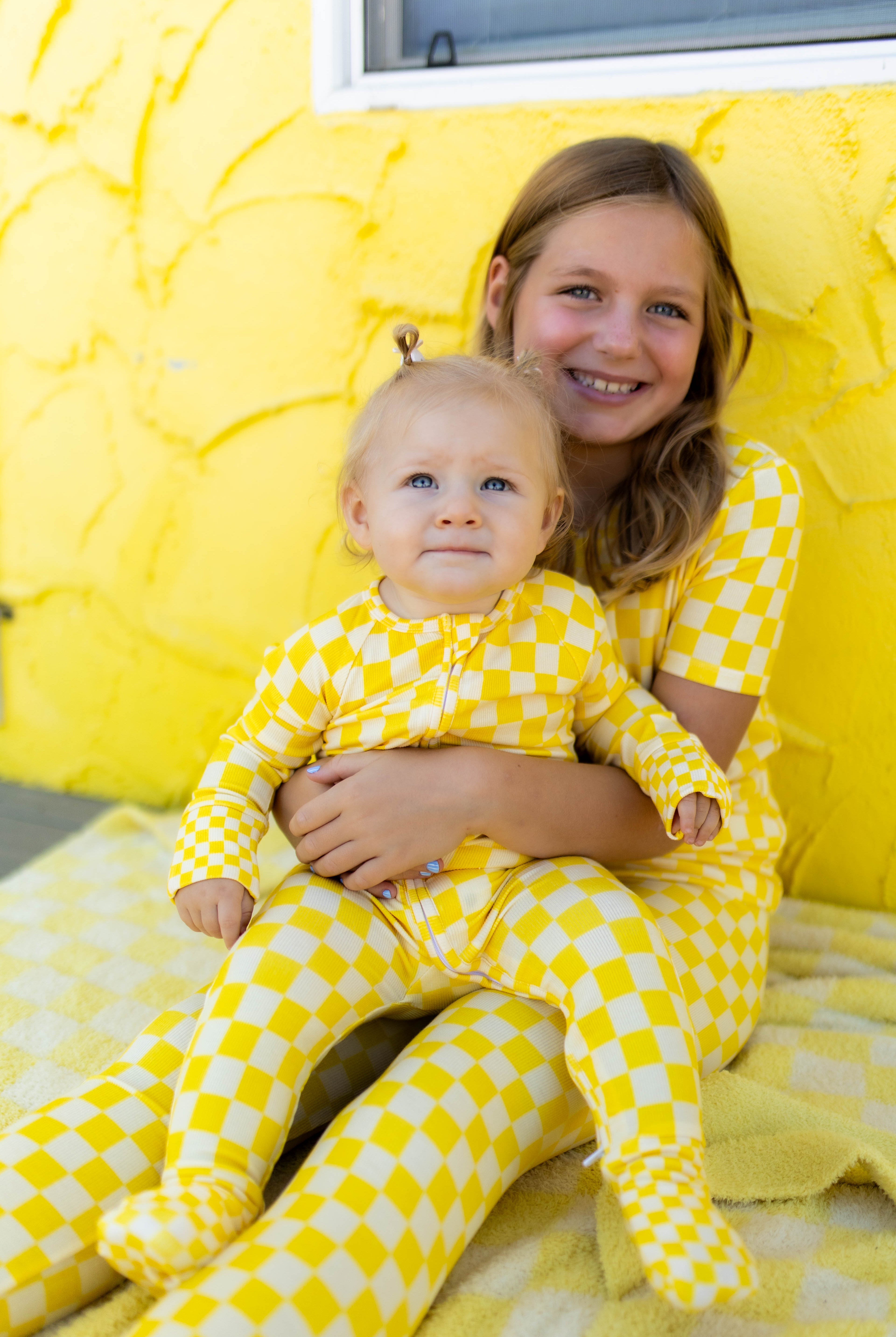 A smiling girl and a baby both wearing matching yellow checkered pajamas, posed together against a bright yellow textured wall.