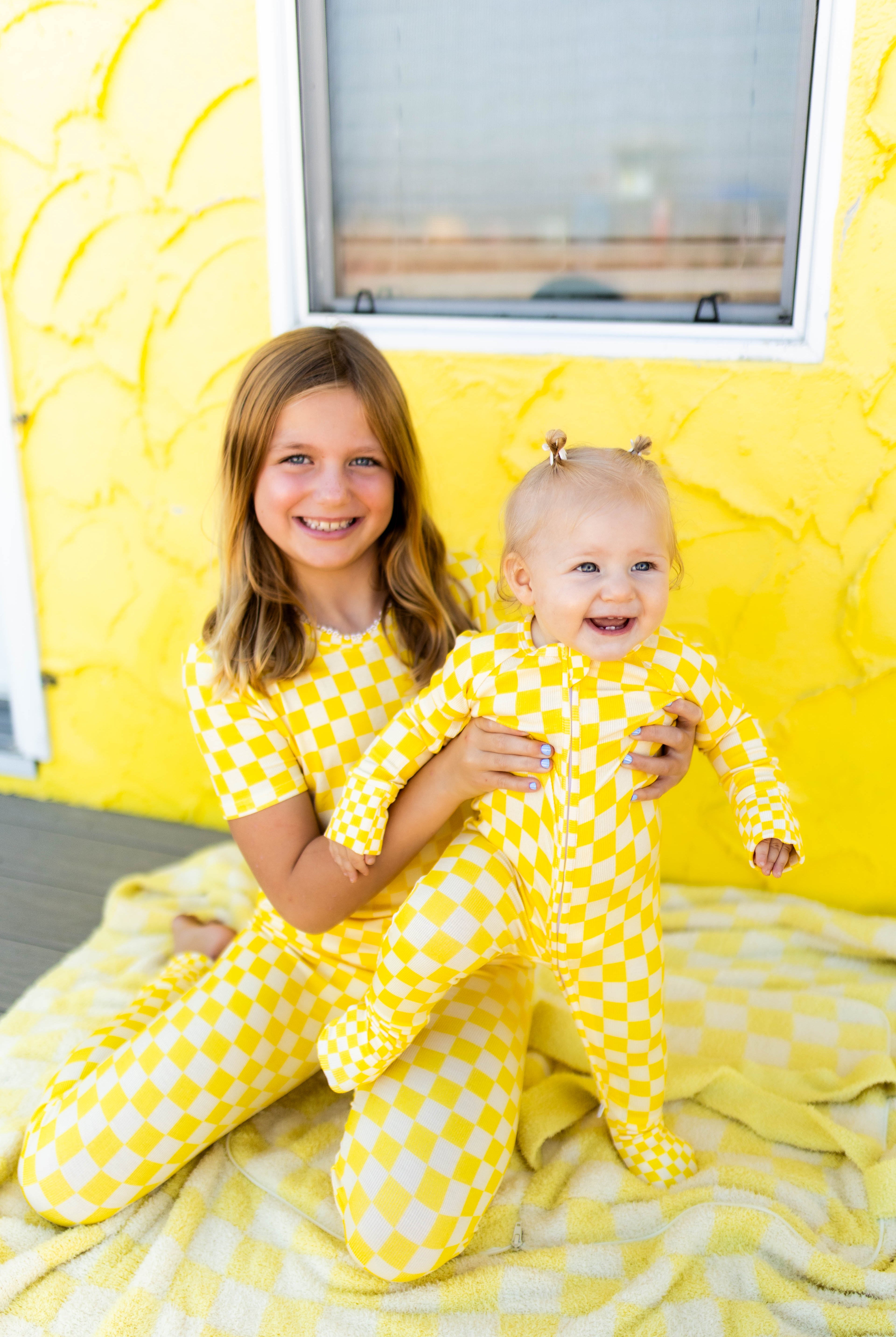 A smiling young girl and a baby sitting together on a yellow blanket in front of a textured yellow wall. Both are wearing matching yellow checkered footed pajamas.