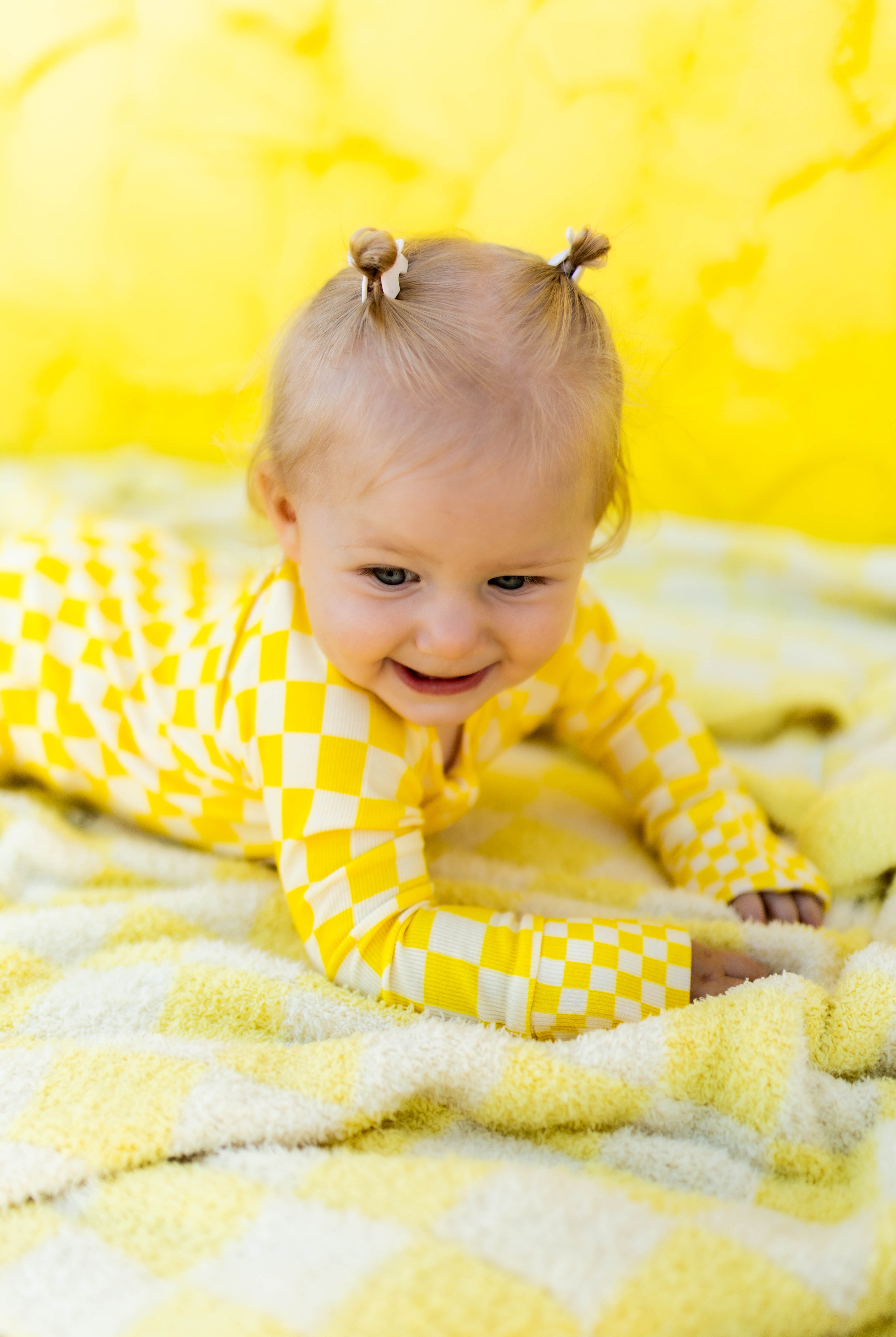 A baby girl wearing a yellow checkered long-sleeve romper, crawling on a matching yellow blanket against a bright yellow background.