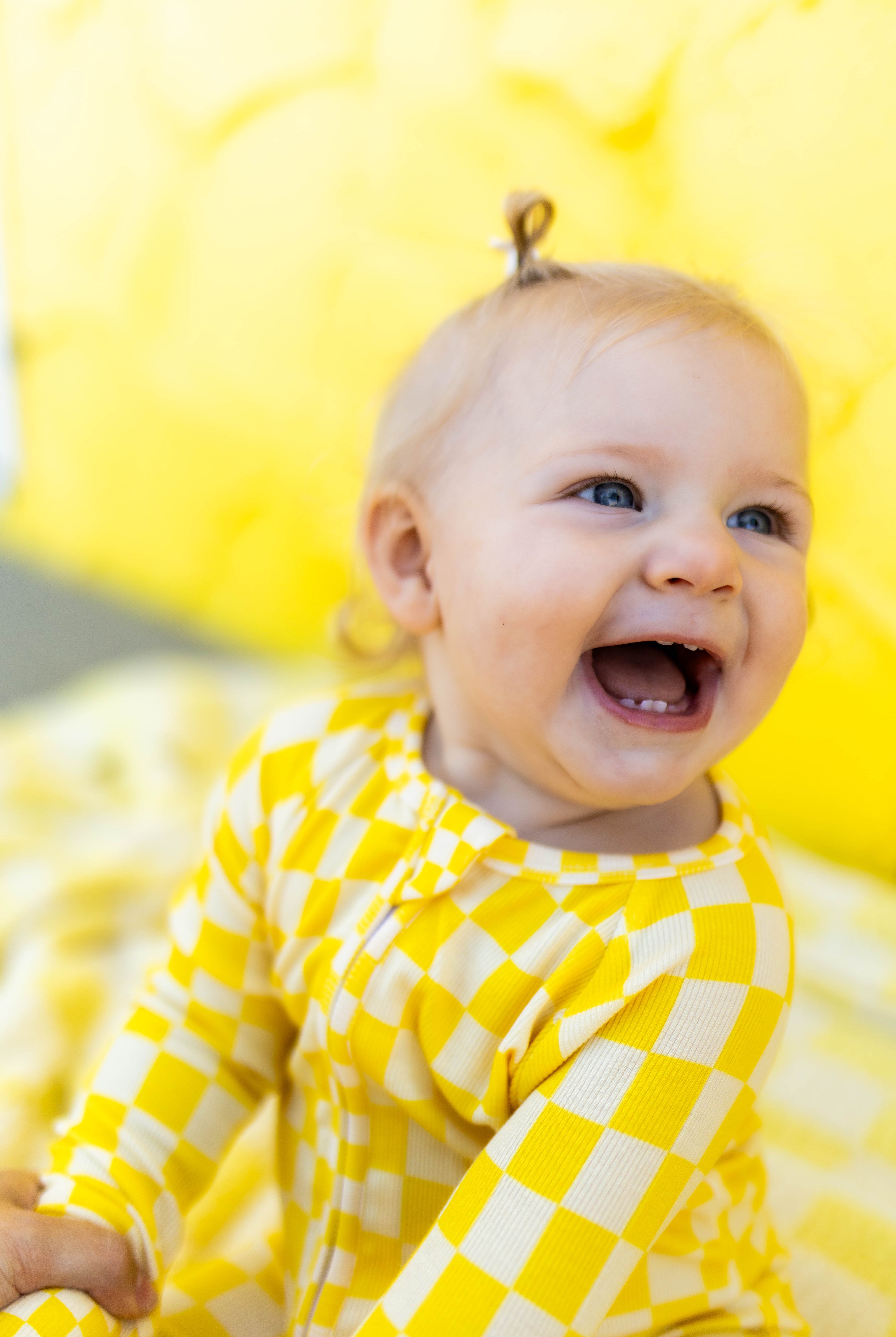 A baby wearing a yellow checkered long-sleeve romper, smiling and laughing while sitting against a bright yellow background.
