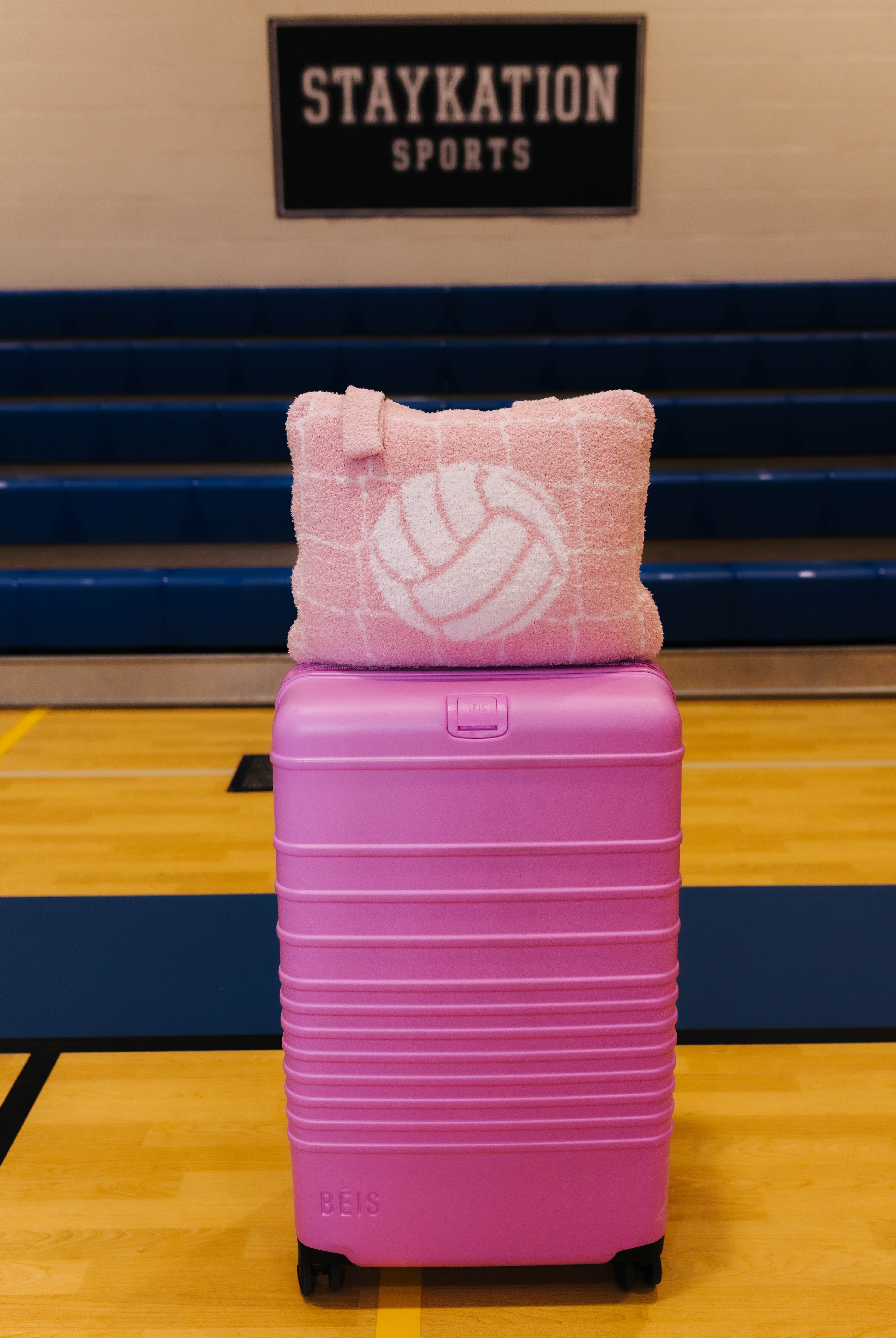 A pink hard-shell suitcase with black wheels sits on a gym floor in front of blue bleachers, with a pink quillow featuring a white volleyball pattern resting on top. A sign reading 'STAYKATION SPORTS' is visible in the background.