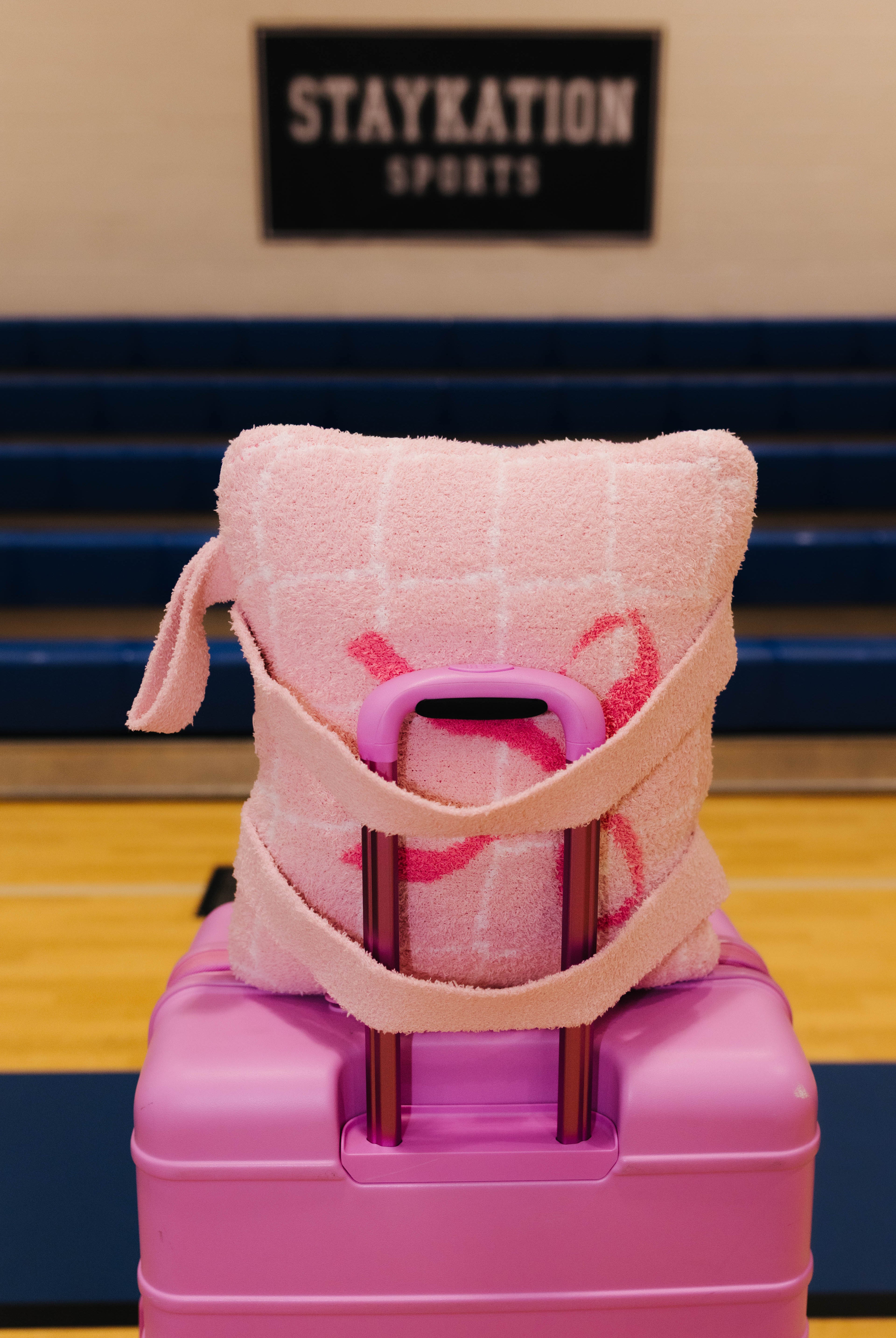 A pink quillow with a grid pattern and pink ribbon accents, wrapped in a matching pink towel strap, resting on top of a pink rolling suitcase in a gymnasium setting.