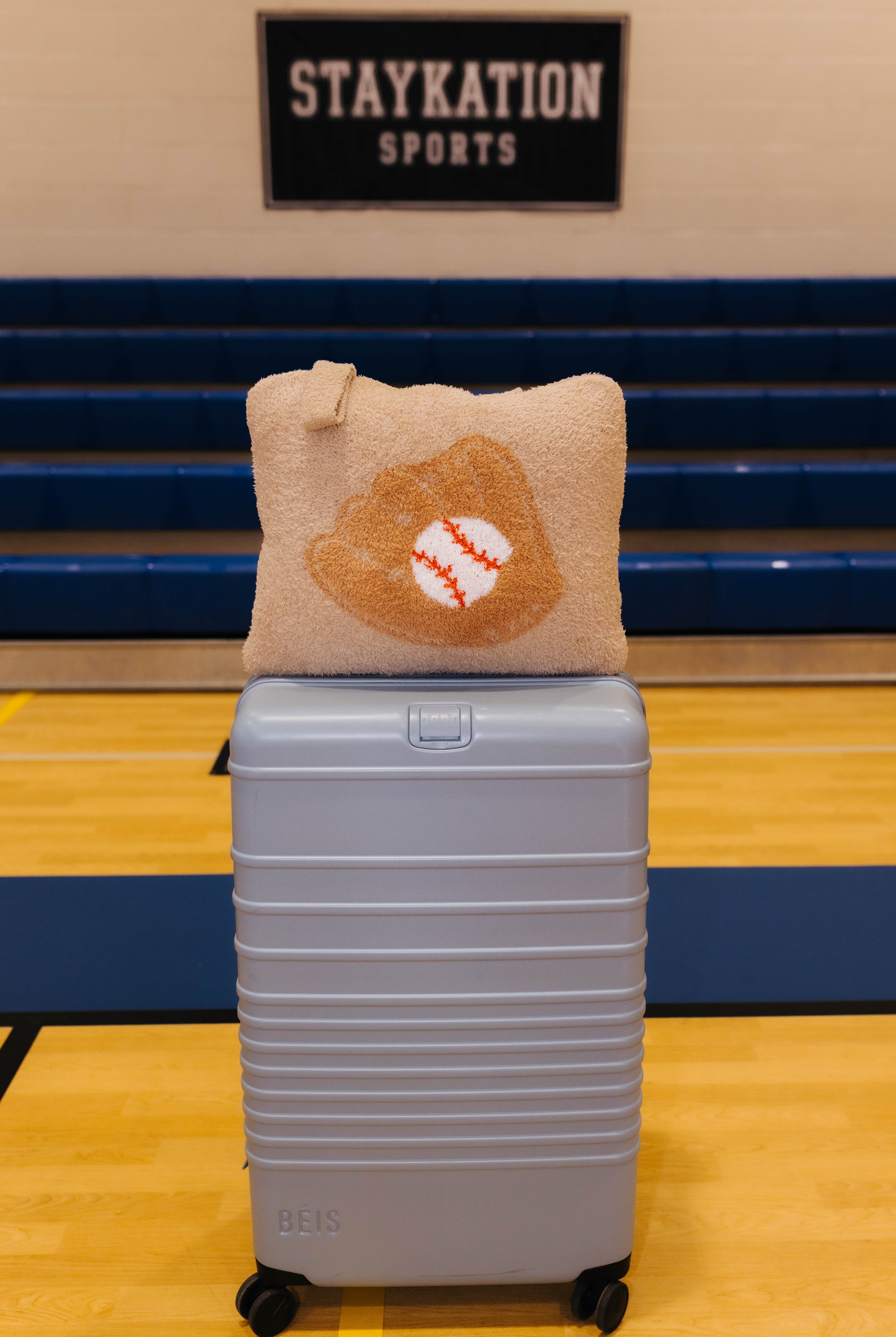 A light blue hard-shell rolling suitcase with a baseball quillow on top it has beige fabric and features a baseball glove design with a baseball in the center.