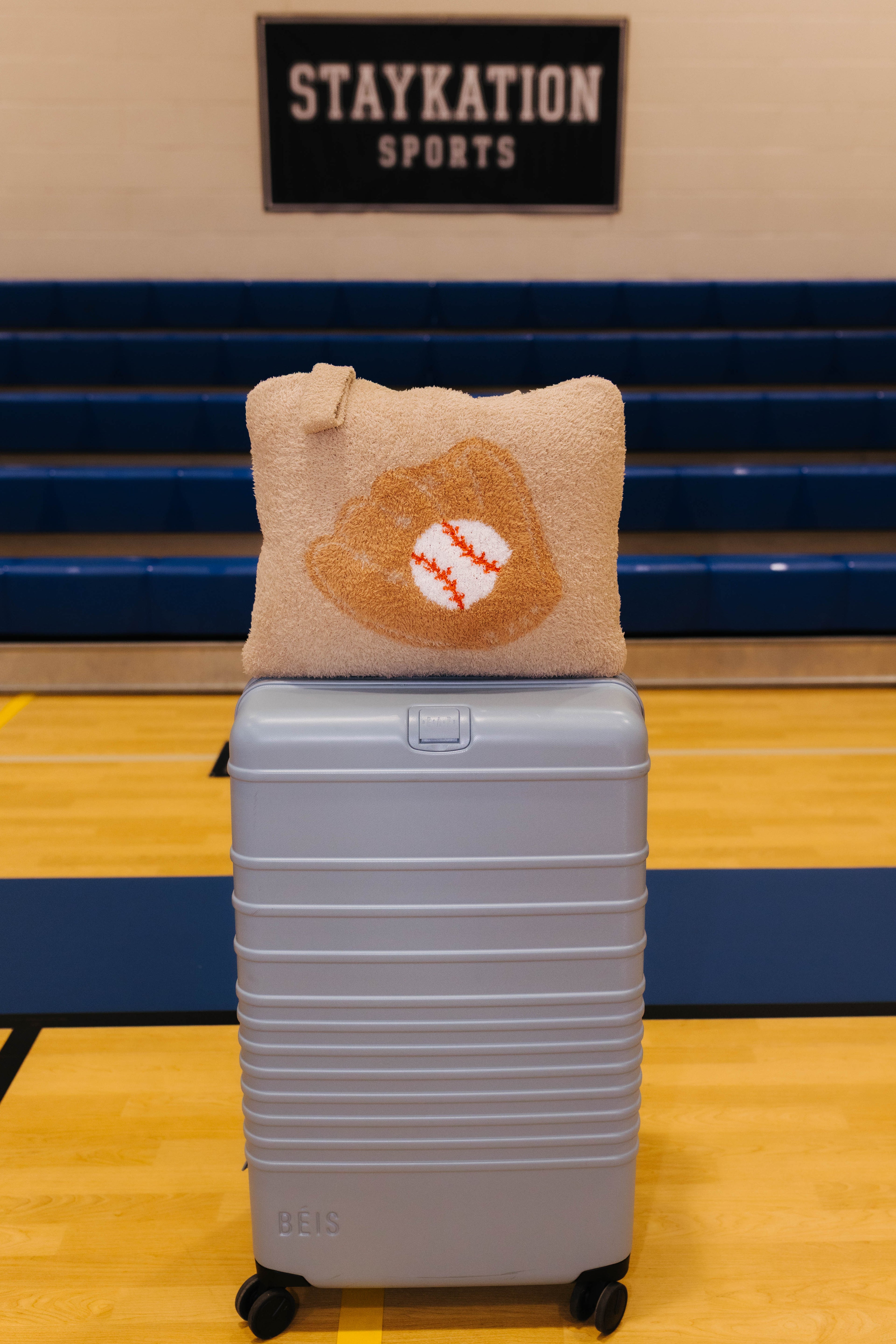 A light blue hard-shell rolling suitcase with a baseball quillow on top it has beige fabric and features a baseball glove design with a baseball in the center.