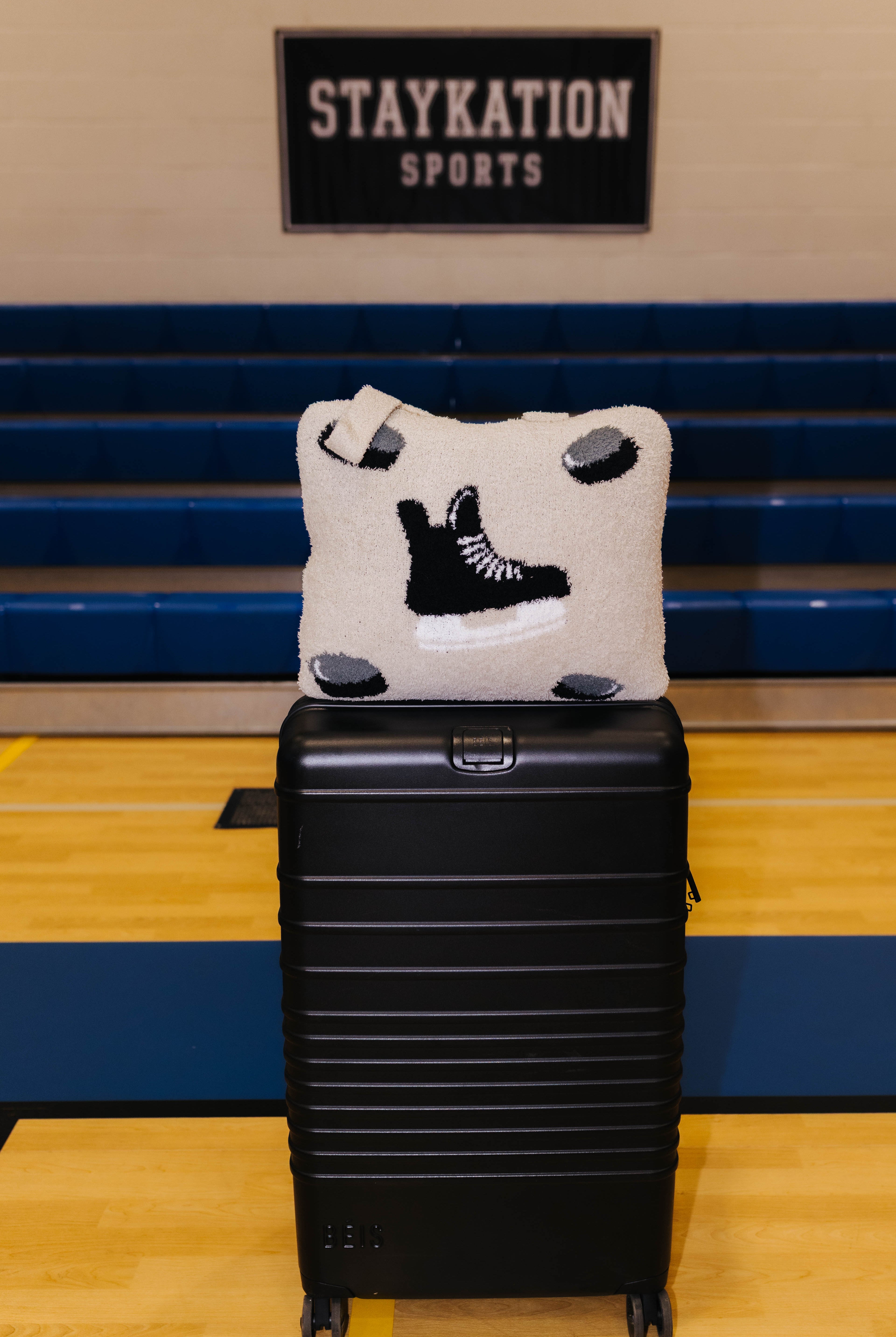 A black hard-shell suitcase standing on a wooden gym floor with blue bleachers and a wall sign in the background. On top of the suitcase is a quillow featuring a black hockey skate design.