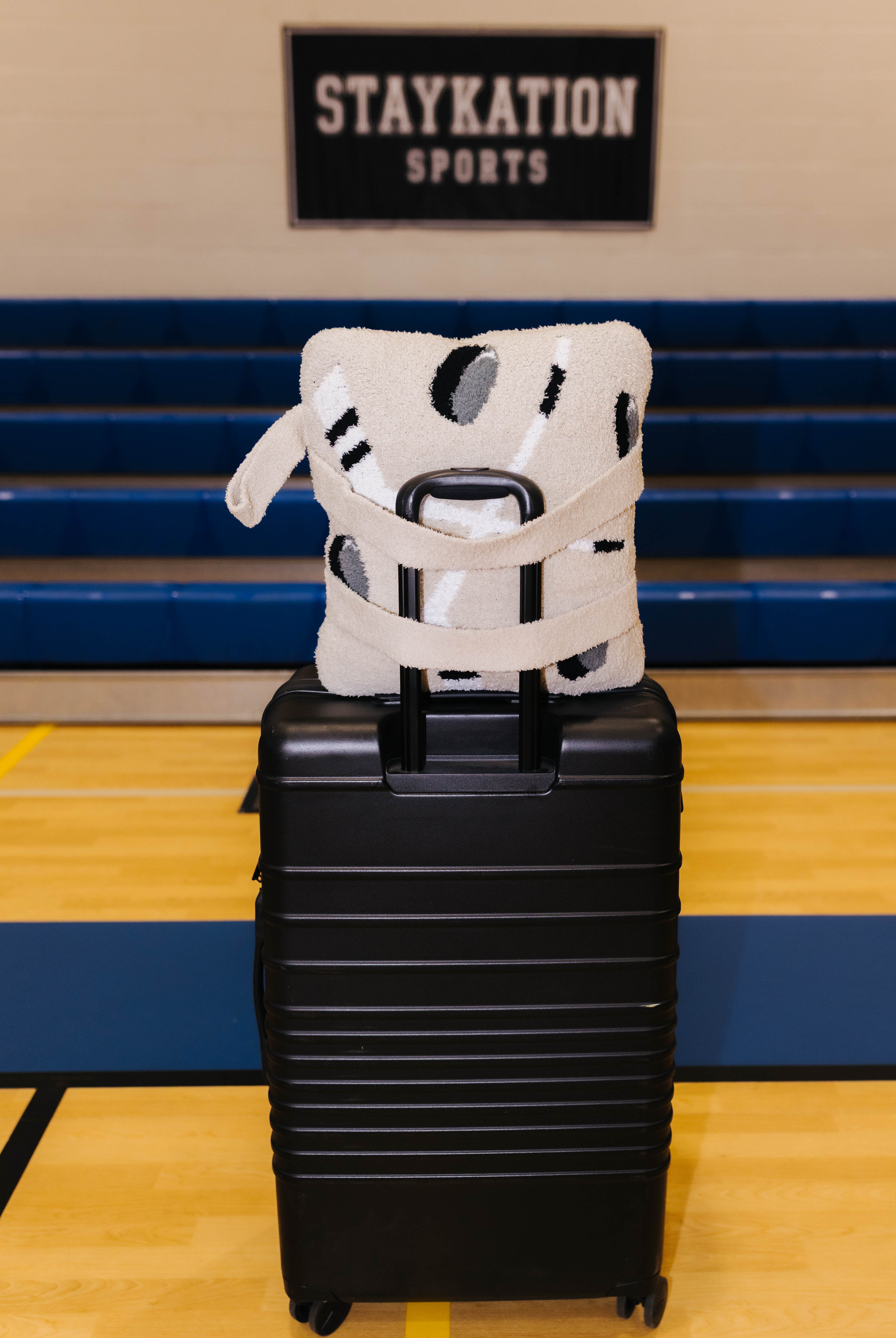A black hard-shell rolling suitcase stands on a gym floor with a white and black patterned hockey quillow on top. Blue bleachers and a wall sign reading STAYKATION SPORTS are visible in the background.