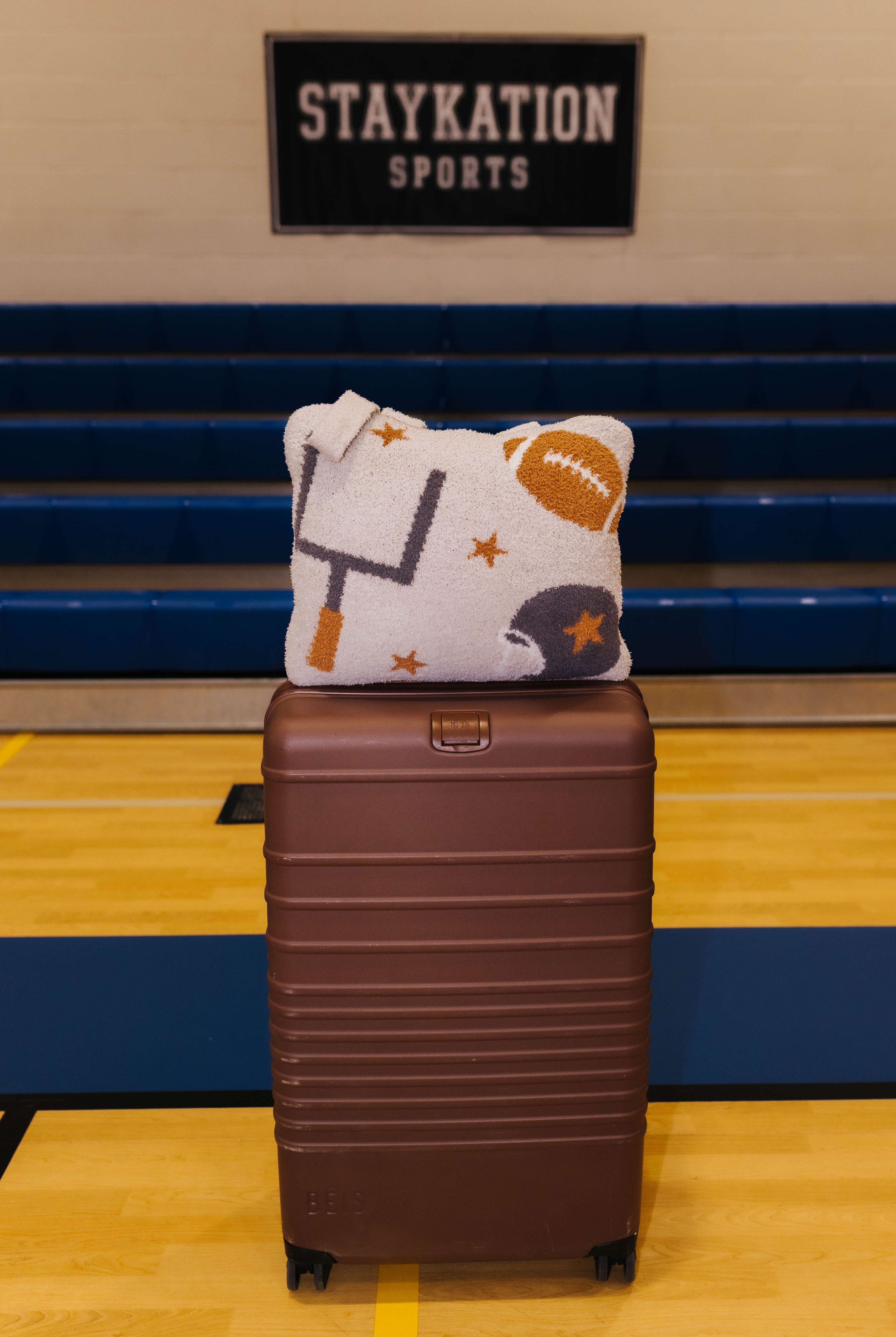 A brown hard-shell rolling suitcase stands on a gym floor in front of blue bleachers, with a tan quillow featuring football-themed graphics resting on top. A 'STAYKATION SPORTS' sign is visible on the wall behind.