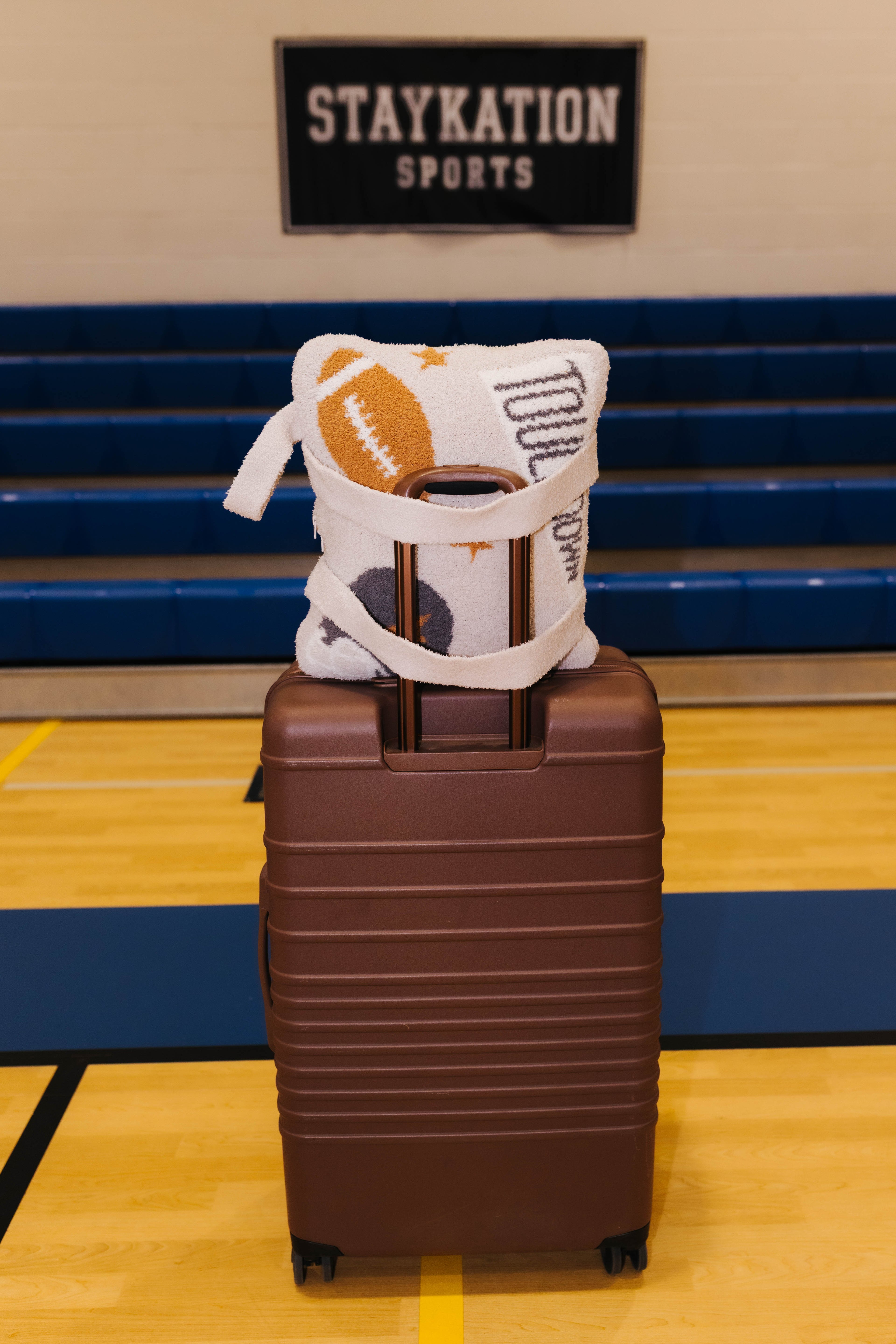 A brown hard-shell suitcase standing on a gym floor with the tan quillow with a football and printed with text, set against blue bleachers and a sign reading STAYKATION SPORTS.