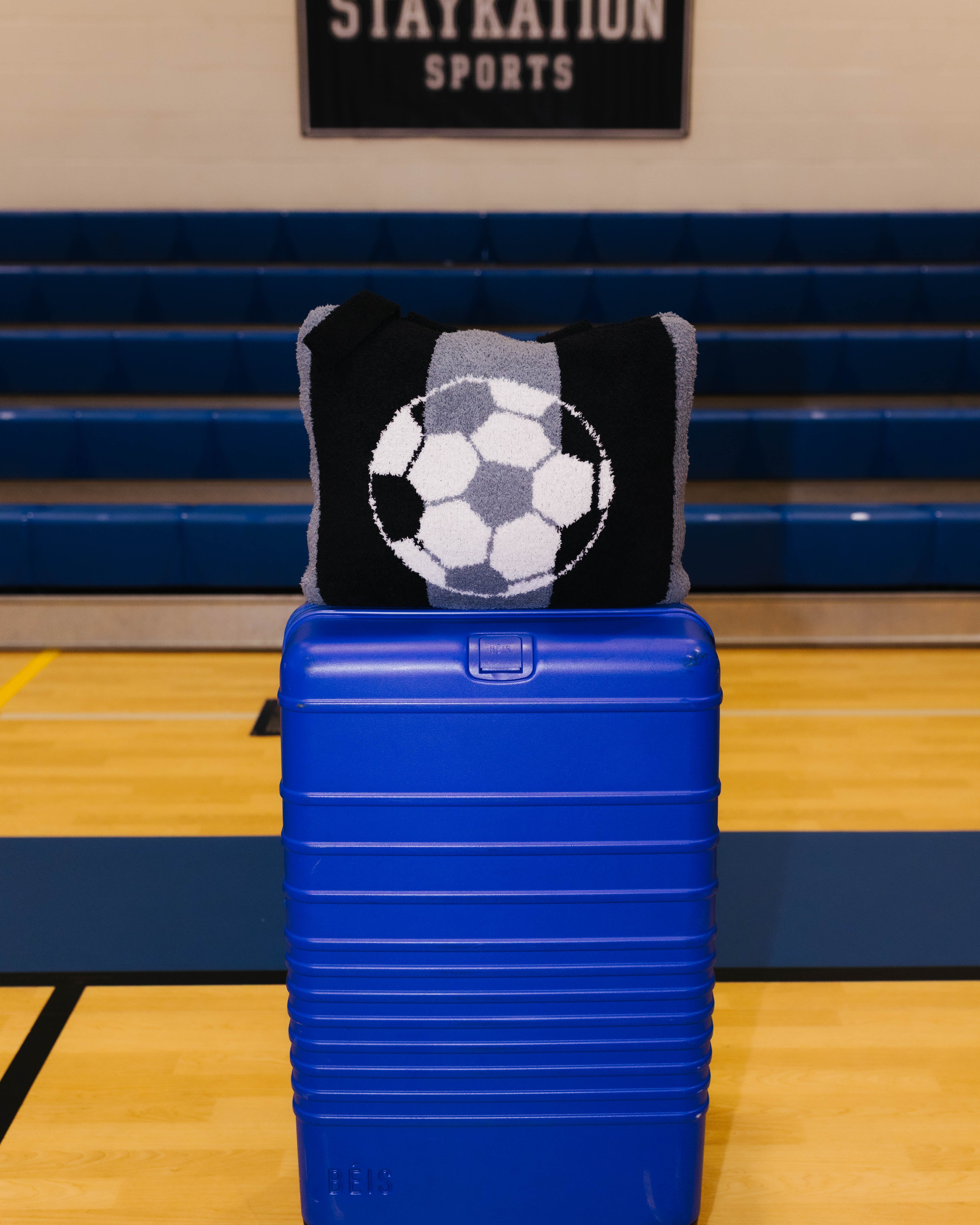 A bright blue hard-shell rolling suitcase stands on a gym floor in front of blue bleachers, with a black and gray knit quillow featuring a soccer ball design resting on top.