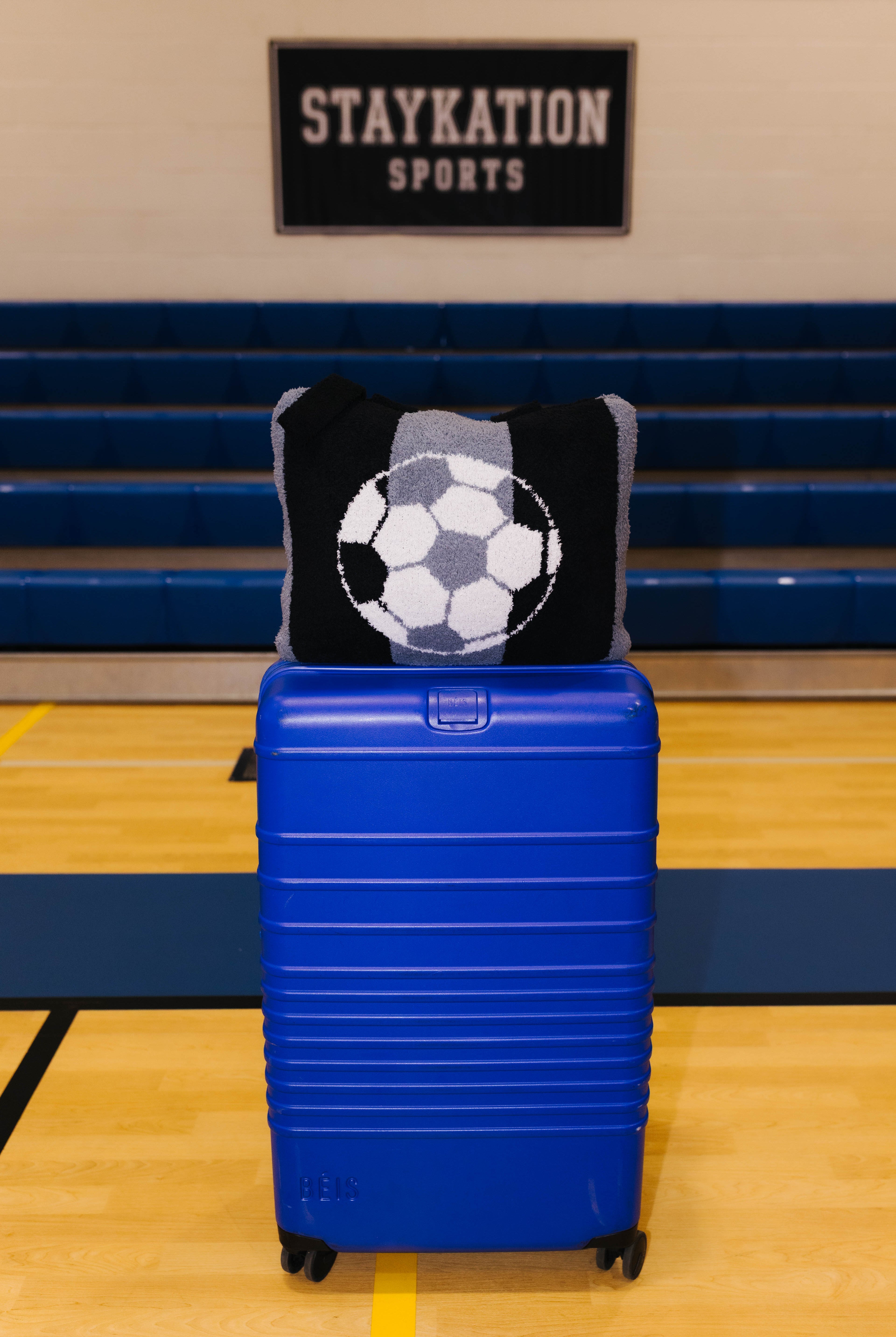A bright blue hard-shell rolling suitcase stands on a gym floor in front of blue bleachers, with a black and gray knit quillow featuring a soccer ball design resting on top.