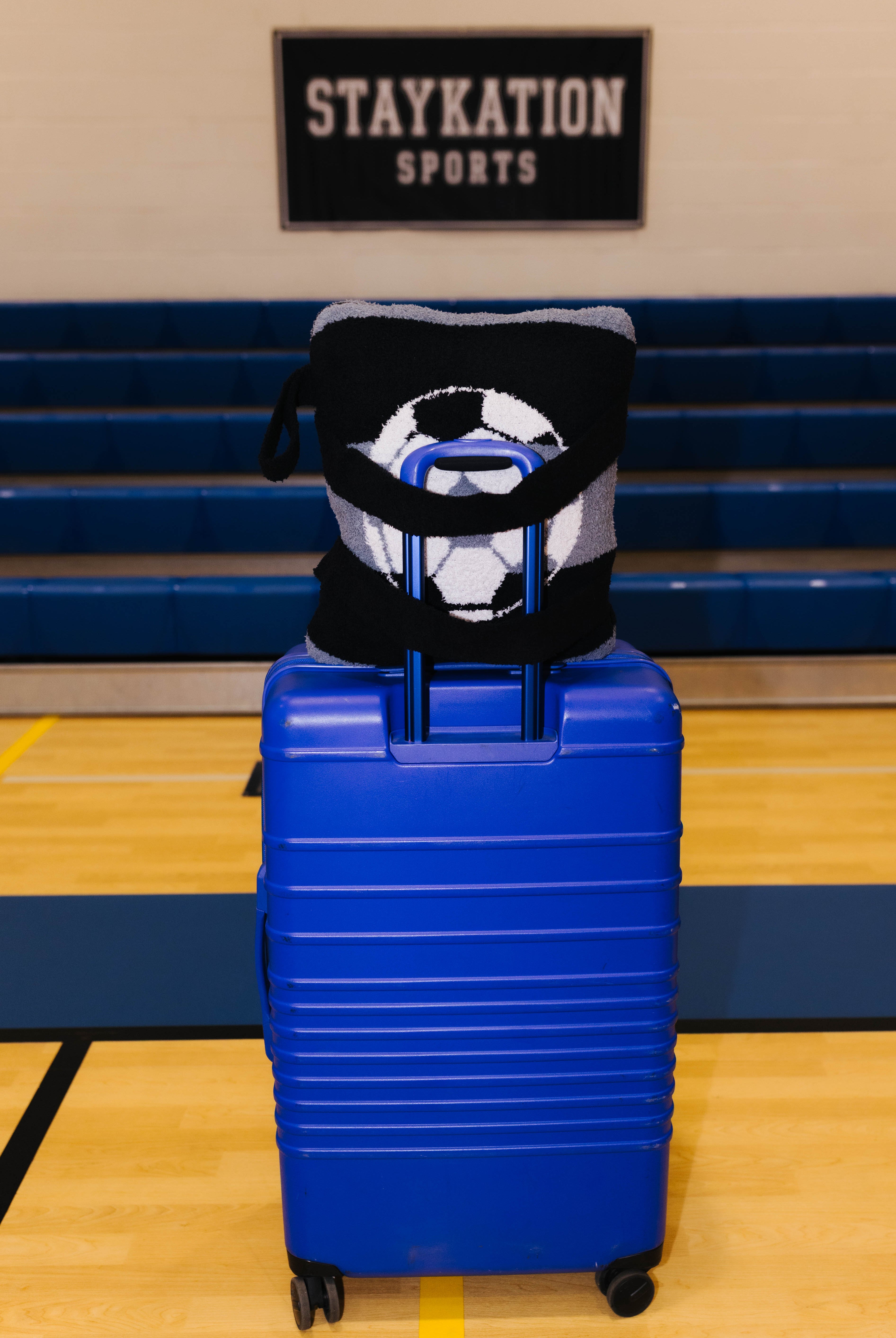 A bright blue hard-shell rolling suitcase stands on an indoor gym floor showing the backside of the quillow.
