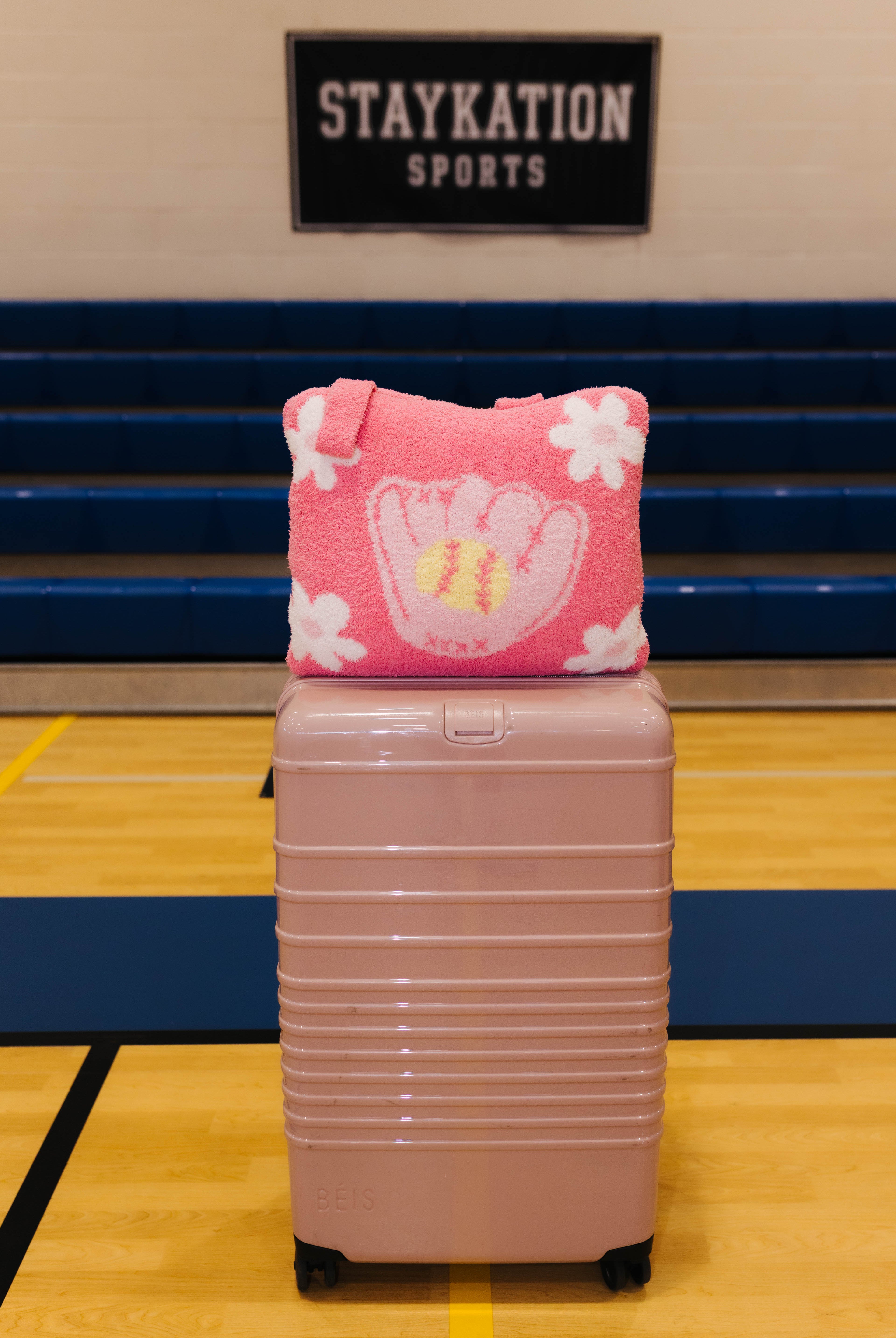 A pink quillow white baseball glove and flower pattern sits on top of a dusty pink hard-shell suitcase in a gymnasium. A 'STAYKATION SPORTS' sign is visible in the background.