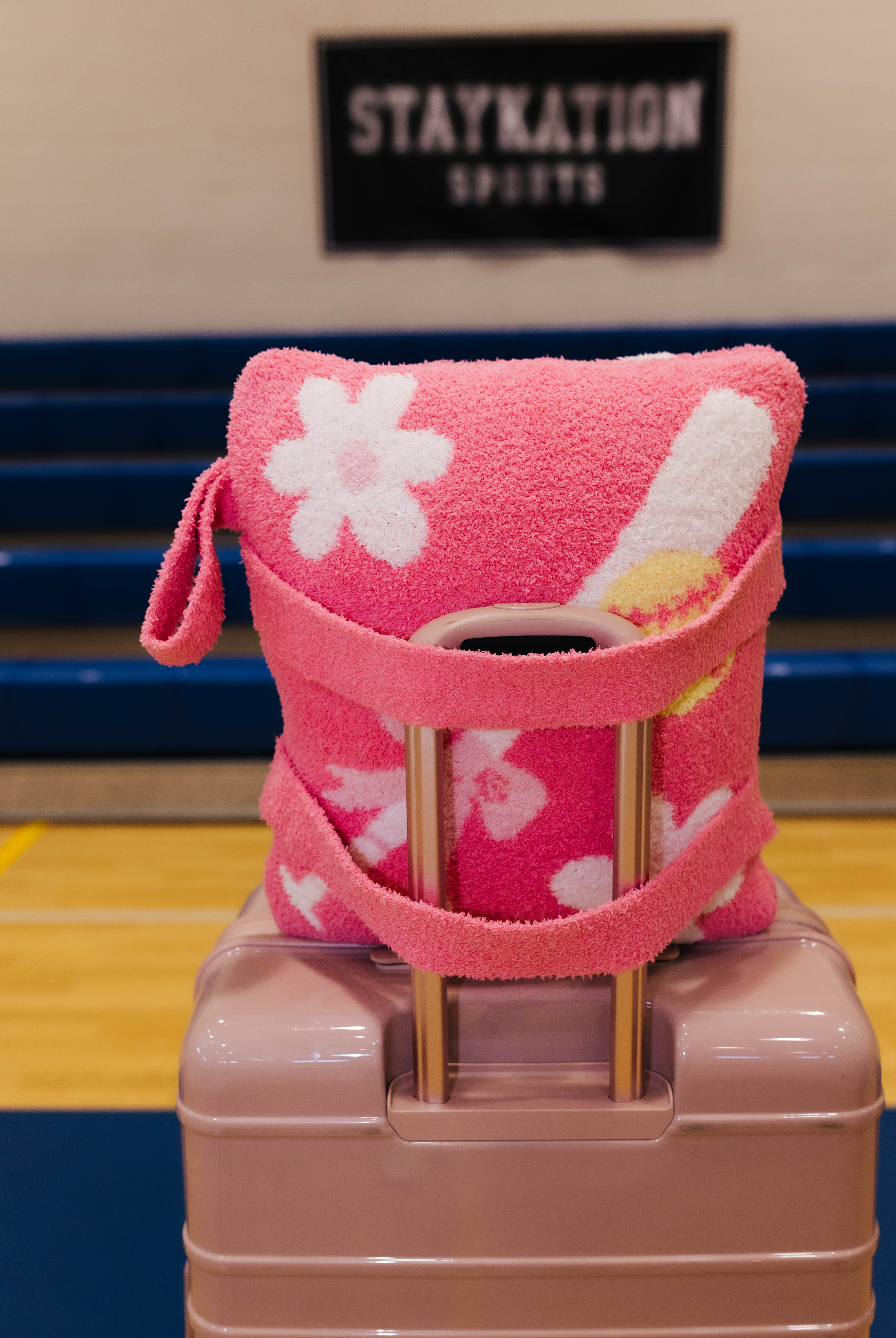 A pink quillow white floral patterns and a pink strap, wrapped around a light pink suitcase in a gym setting with a 'STAYKATION SPORTS' sign in the background.