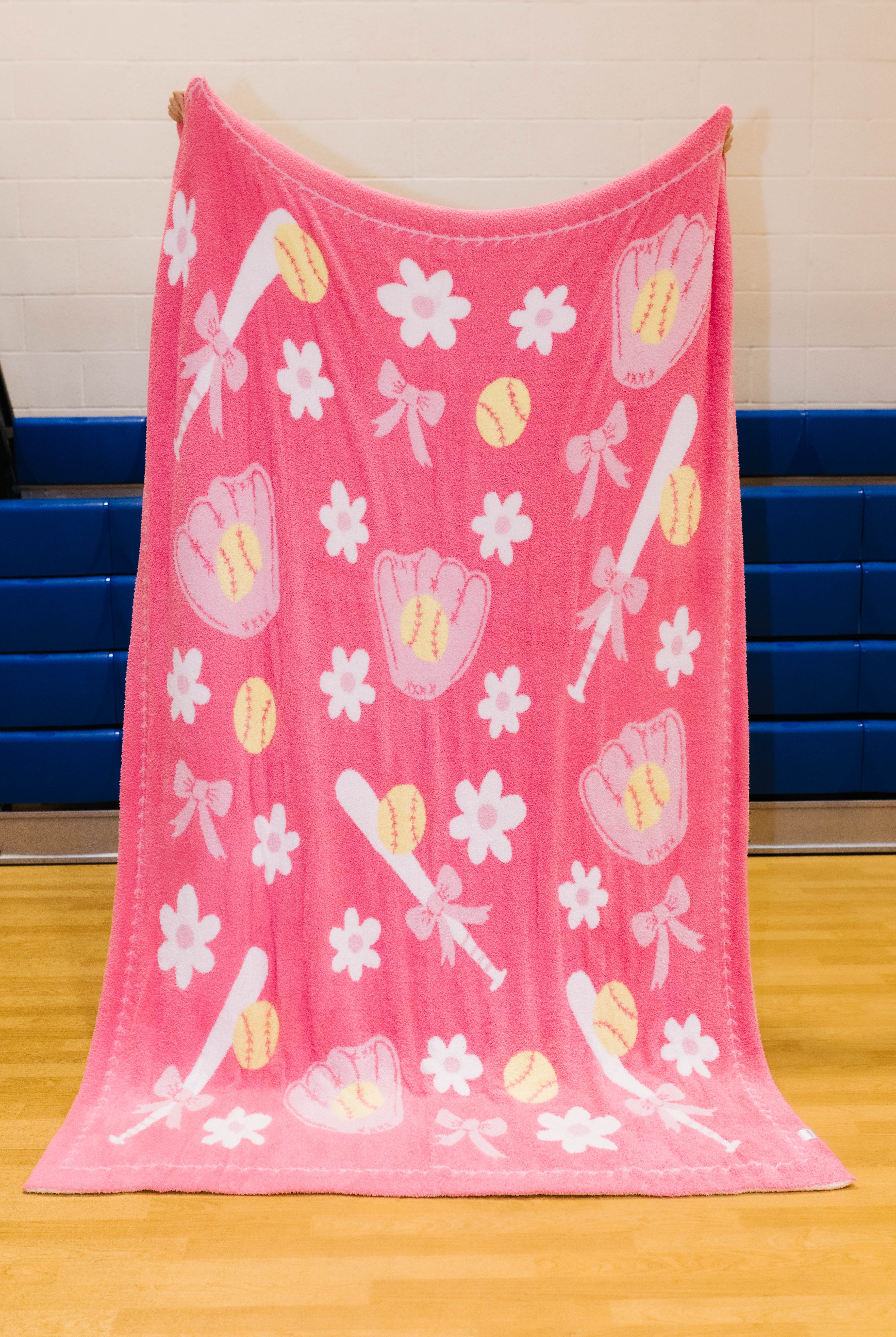 A pink fleece blanket with a baseball and softball theme, featuring white flowers, pink baseball gloves, yellow softballs, and white bats, hanging on a gym wall.