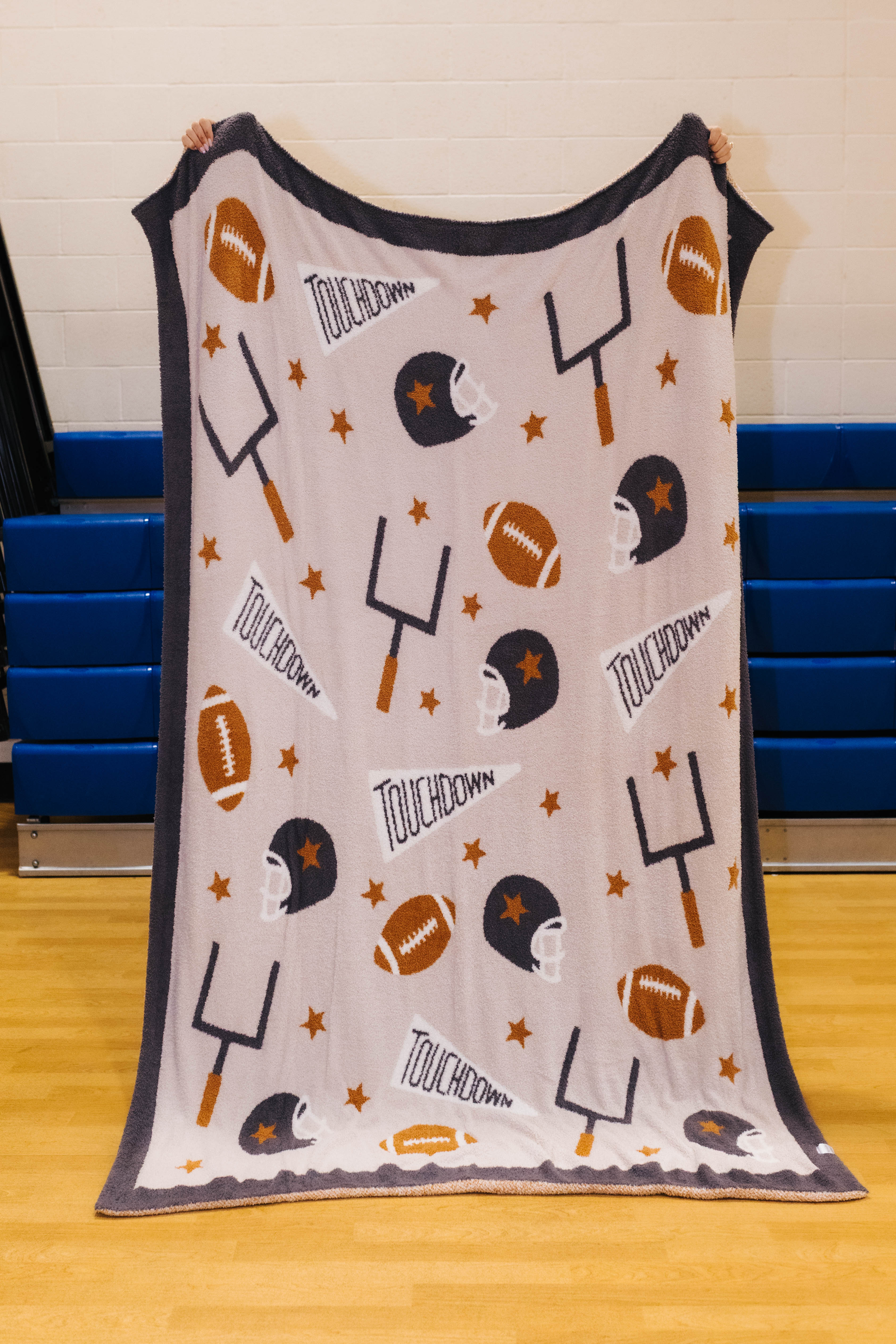 A throw blanket featuring a football theme with 'TOUCHDOWN' text, footballs, helmets, and turf icons, held up in a gym setting.