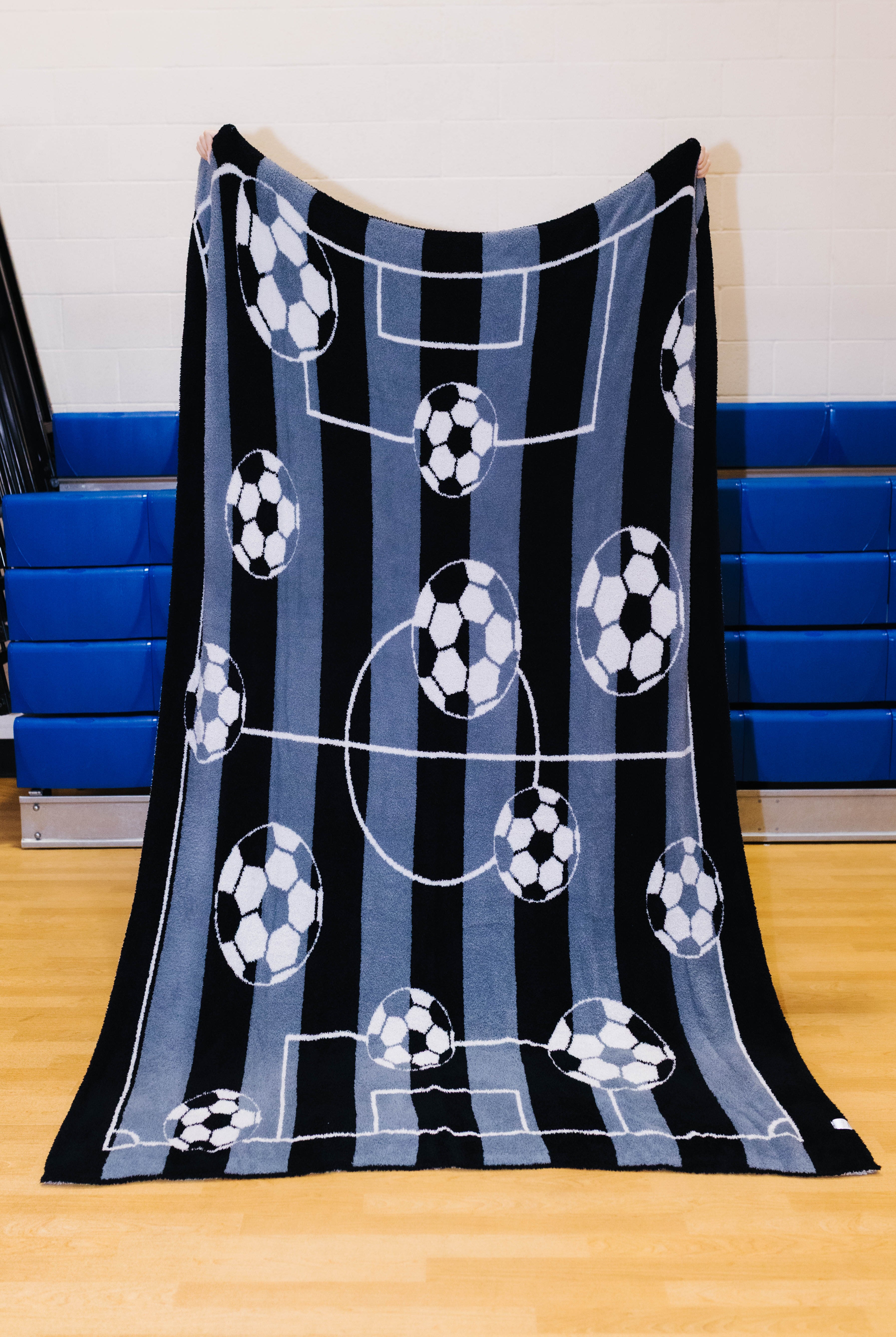 A fleece throw blanket featuring a soccer theme with black and blue vertical stripes, white field lines, and soccer balls, held up by a person in an indoor gym setting.