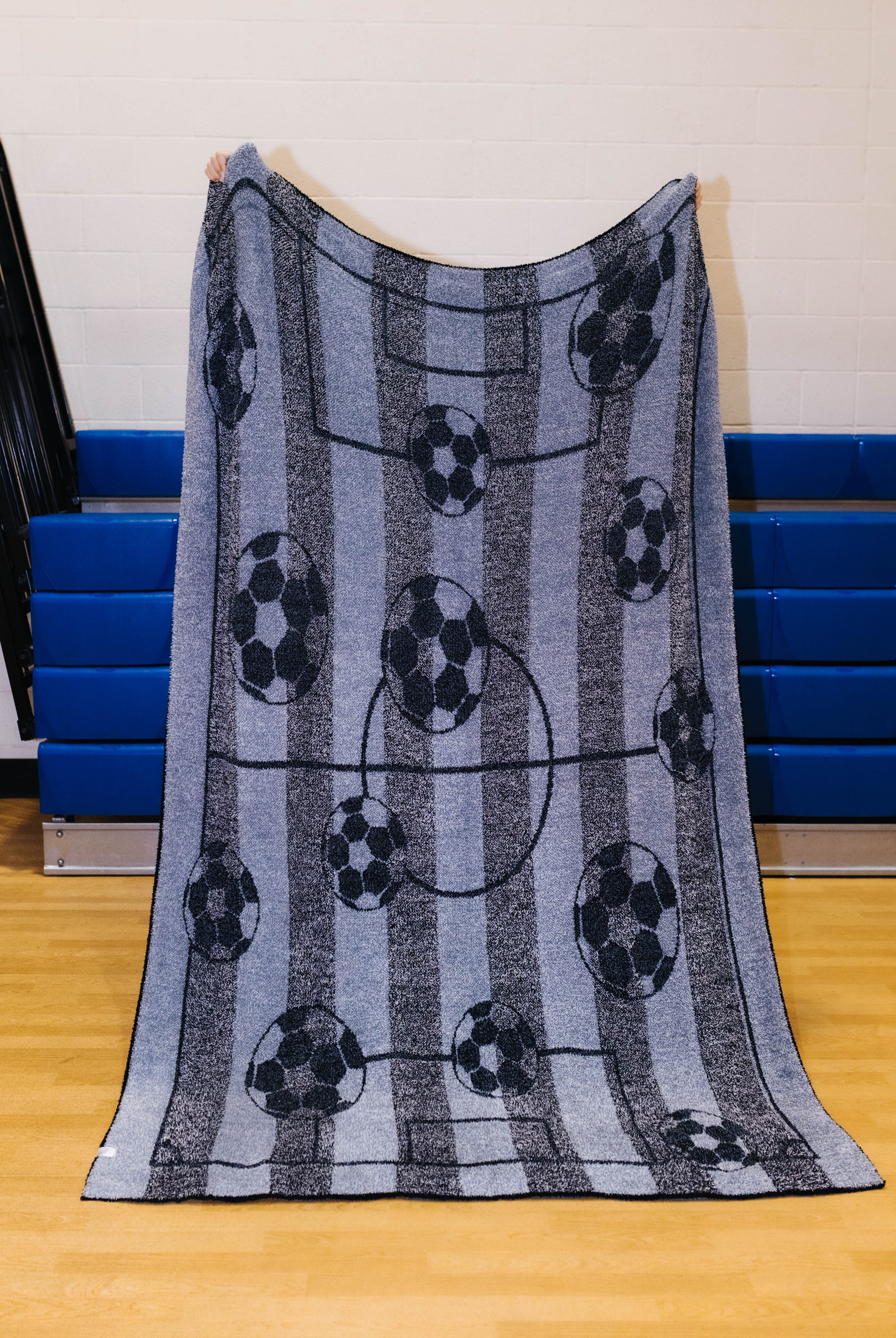 Back side of the rectangular fleece blanket with a soccer theme, featuring black and white soccer balls on vertical gray and blue-gray stripes with a faint soccer field graphic, held up by a person in an indoor gym setting.