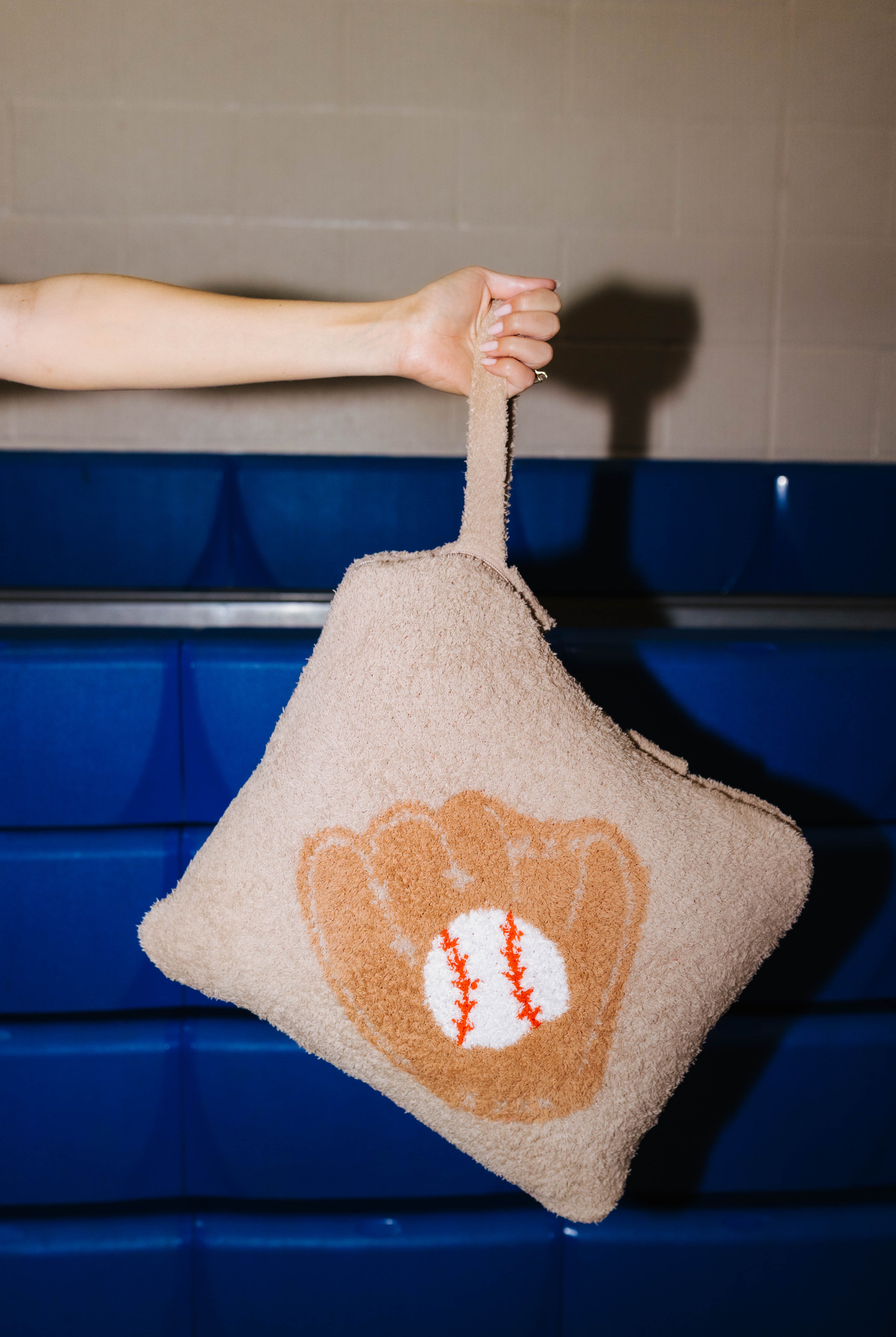 A beige baseball-themed quillow held by a person's arm against a blue padded wall and gray tiled background.