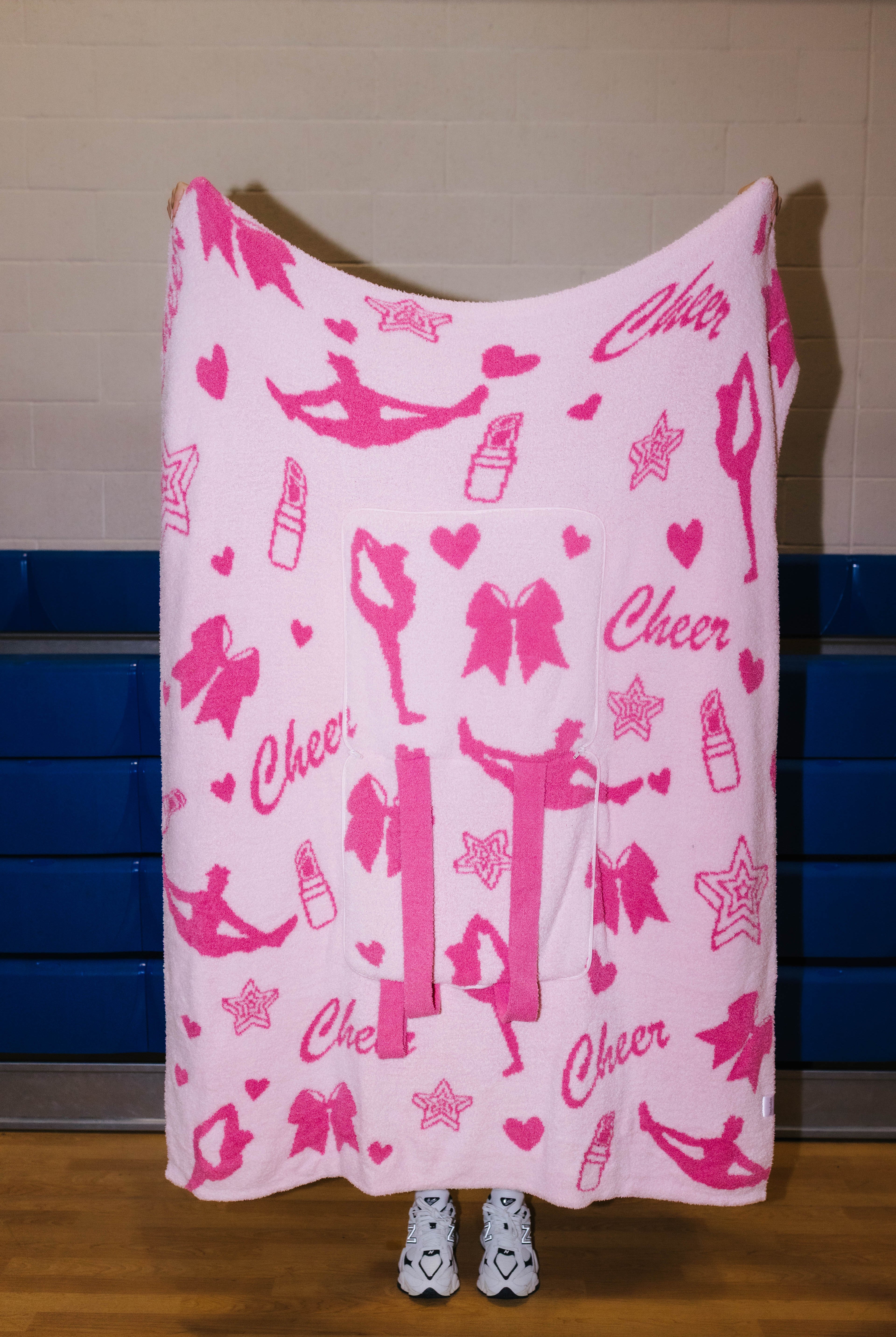 A pink fleece blanket with a pink and white cheerleader-themed print featuring dancers, bows, lipstick tubes, stars, and the word 'Cheer', held up by a person standing in front of a gym wall.