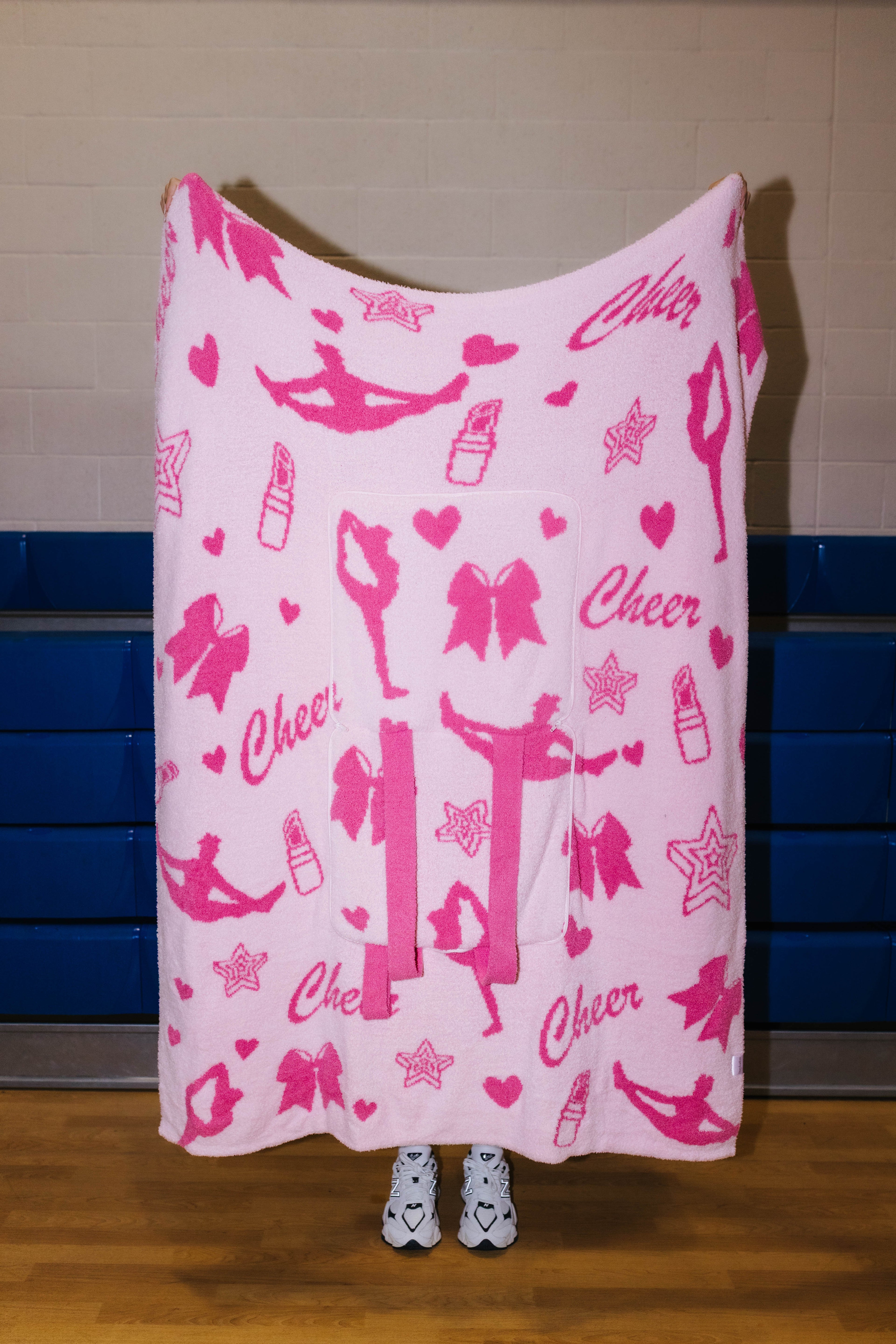 A pink fleece blanket with a pink and white cheerleader-themed print featuring dancers, bows, lipstick tubes, stars, and the word 'Cheer', held up by a person standing in front of a gym wall.