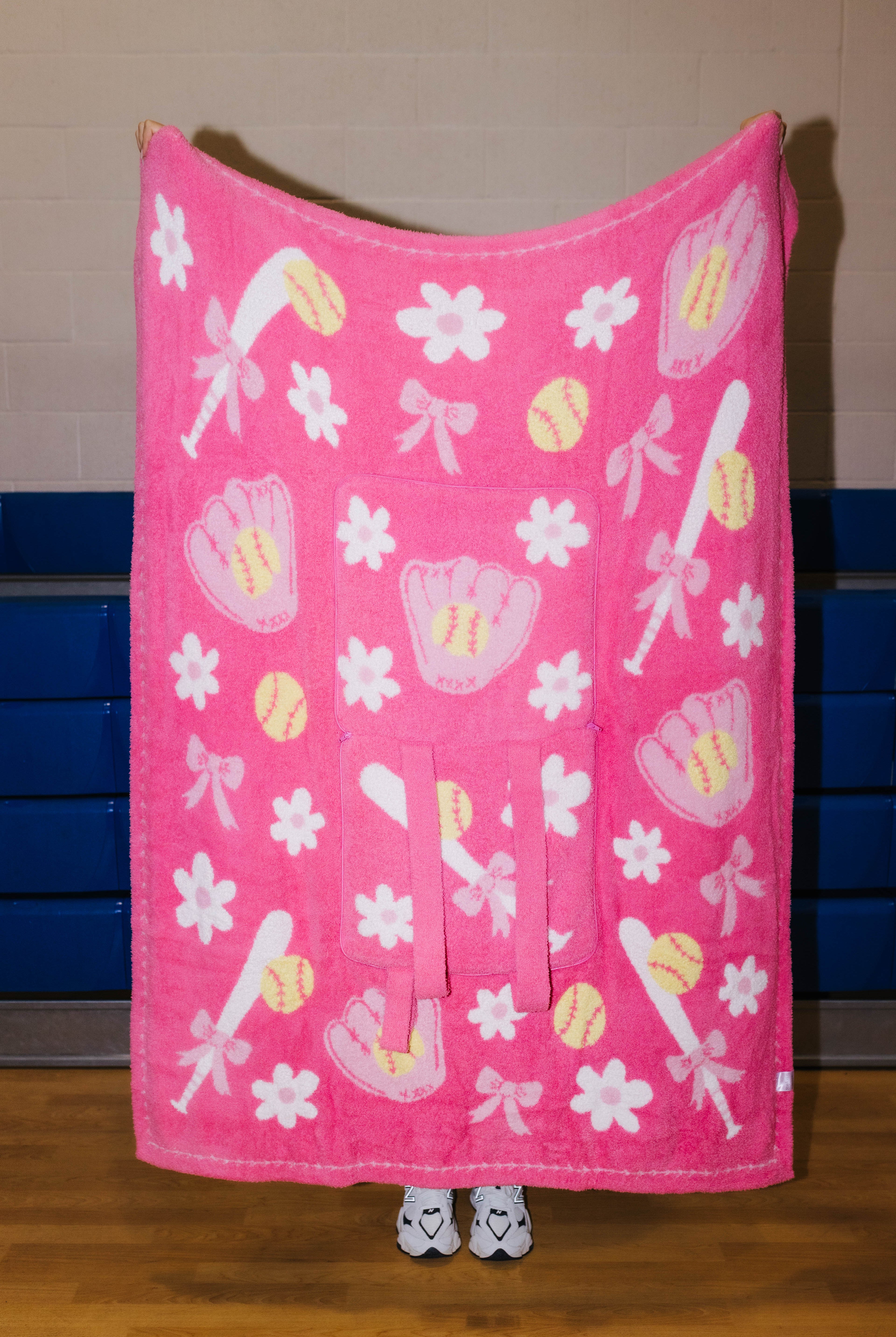 A pink fleece blanket with a baseball and softball pattern, held up by a person whose feet are visible at the bottom. The blanket features white flowers, pink bows, pink baseball gloves, and yellow baseballs and softballs.