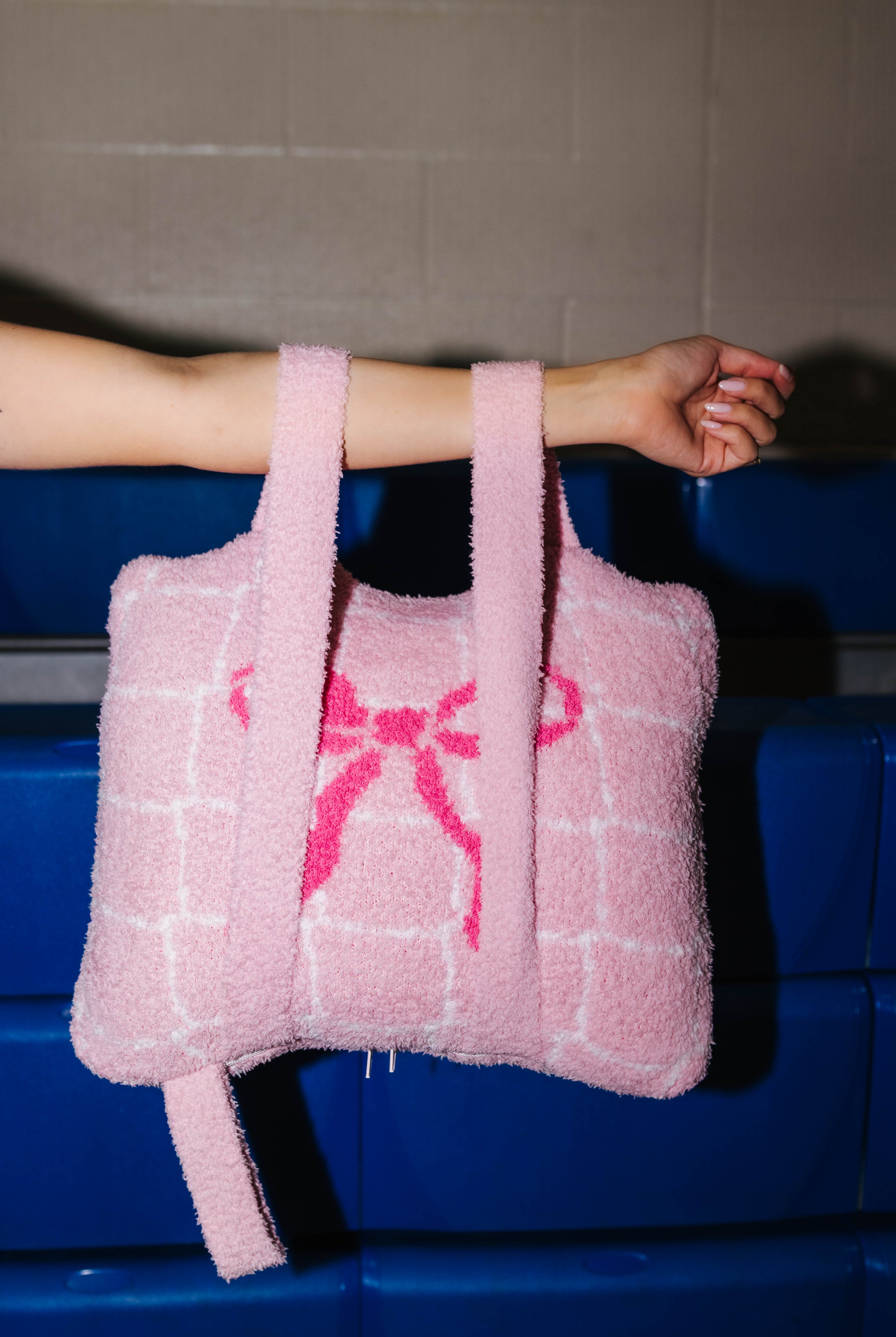 A pink quillow with a grid pattern and a pink bow design, held by a person's arm against a blue bench and gray wall background.