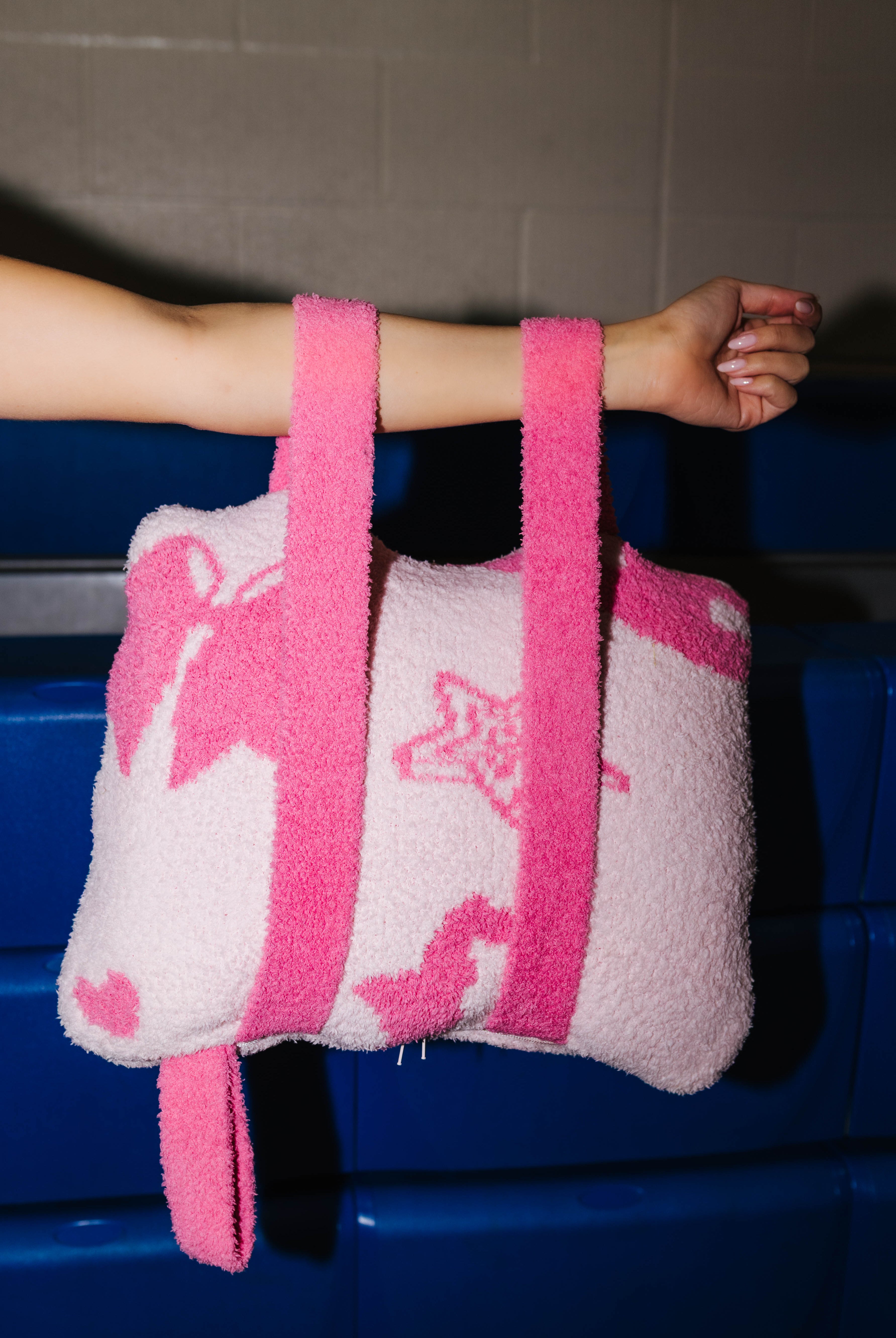 A fuzzy quillow with pink straps, held by a person's arm against a dark blue bench and gray wall background.