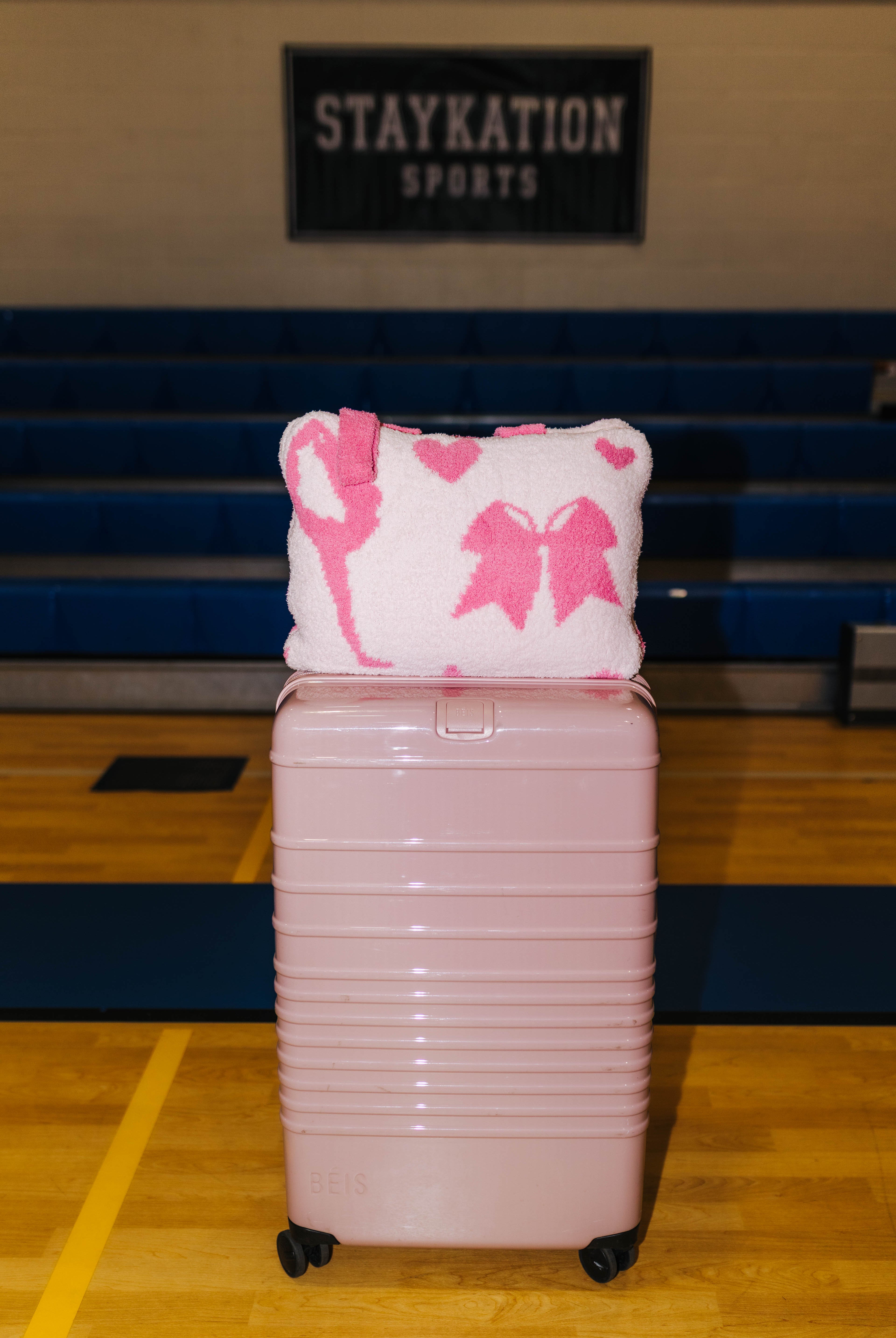 A light pink hard-shell rolling suitcase stands on a wooden gym floor in front of blue bleachers, with a white and pink patterned cheer quillow on top.