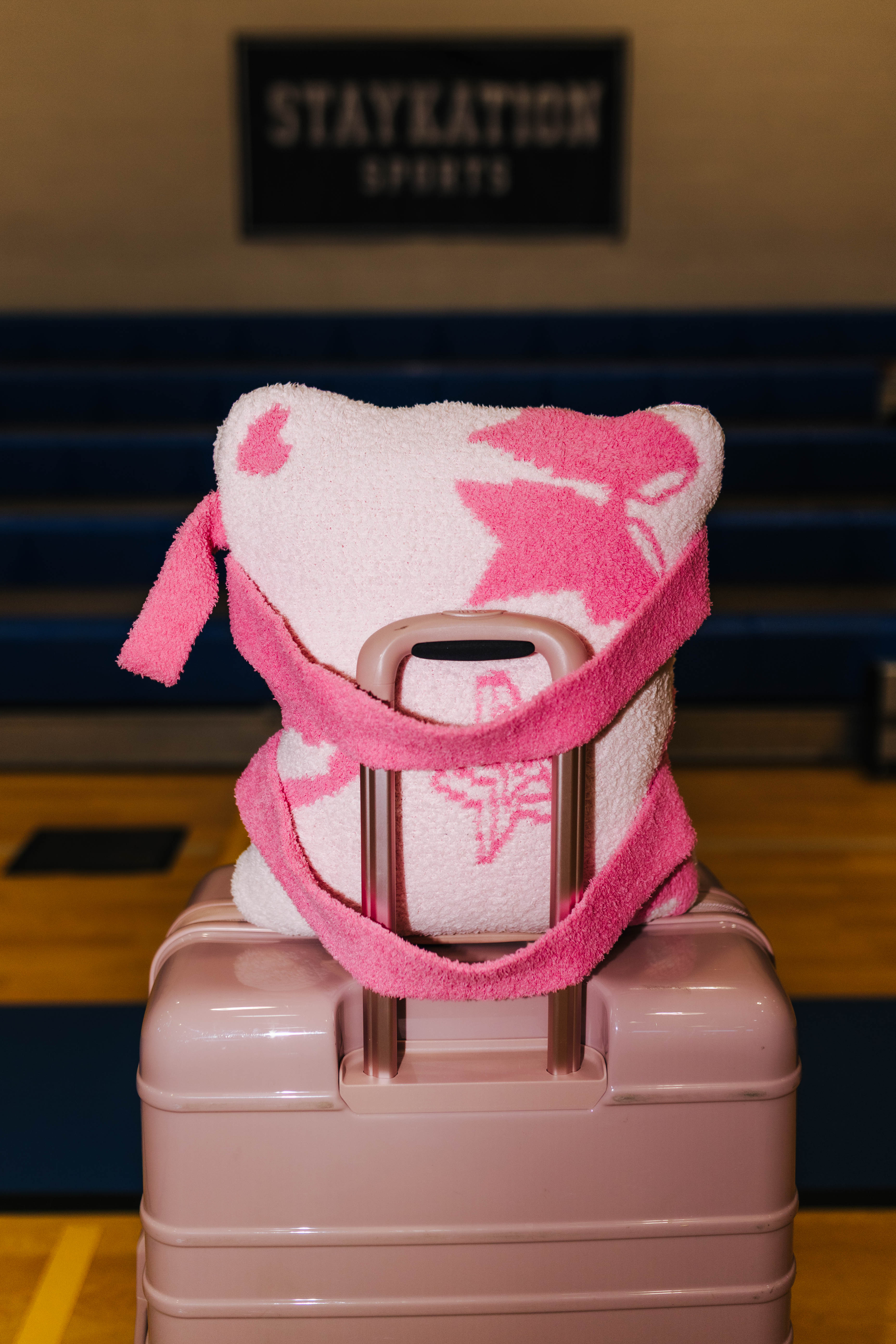 A pink and white patterned quillow on a light pink hard-shell suitcase, set in front of blue stadium seating with a 'STAYKATION SPORTS' sign in the background.
