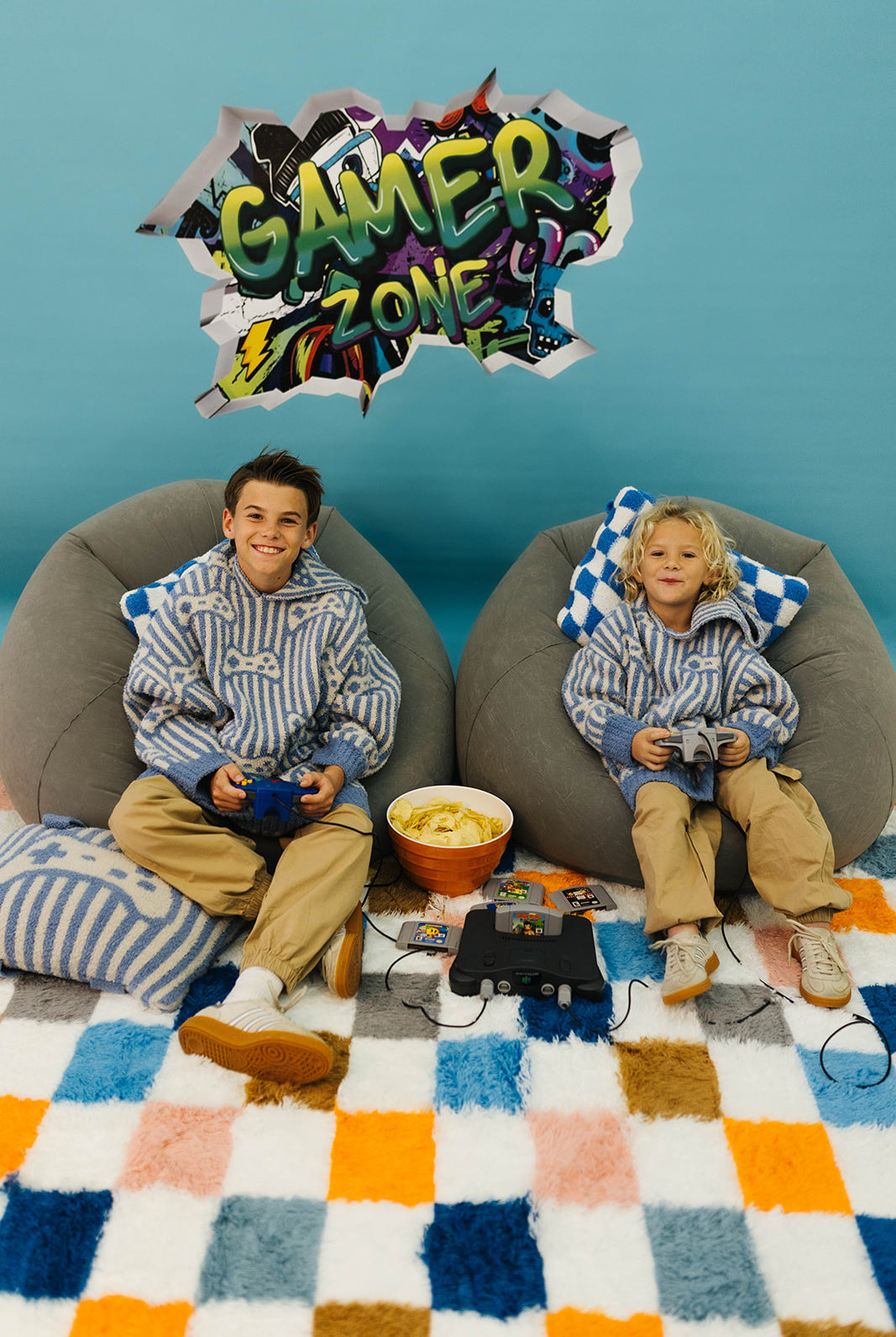 Two children sitting on bean bags with gaming equipment, surrounded by a colorful 'Gamer Zone' sign.