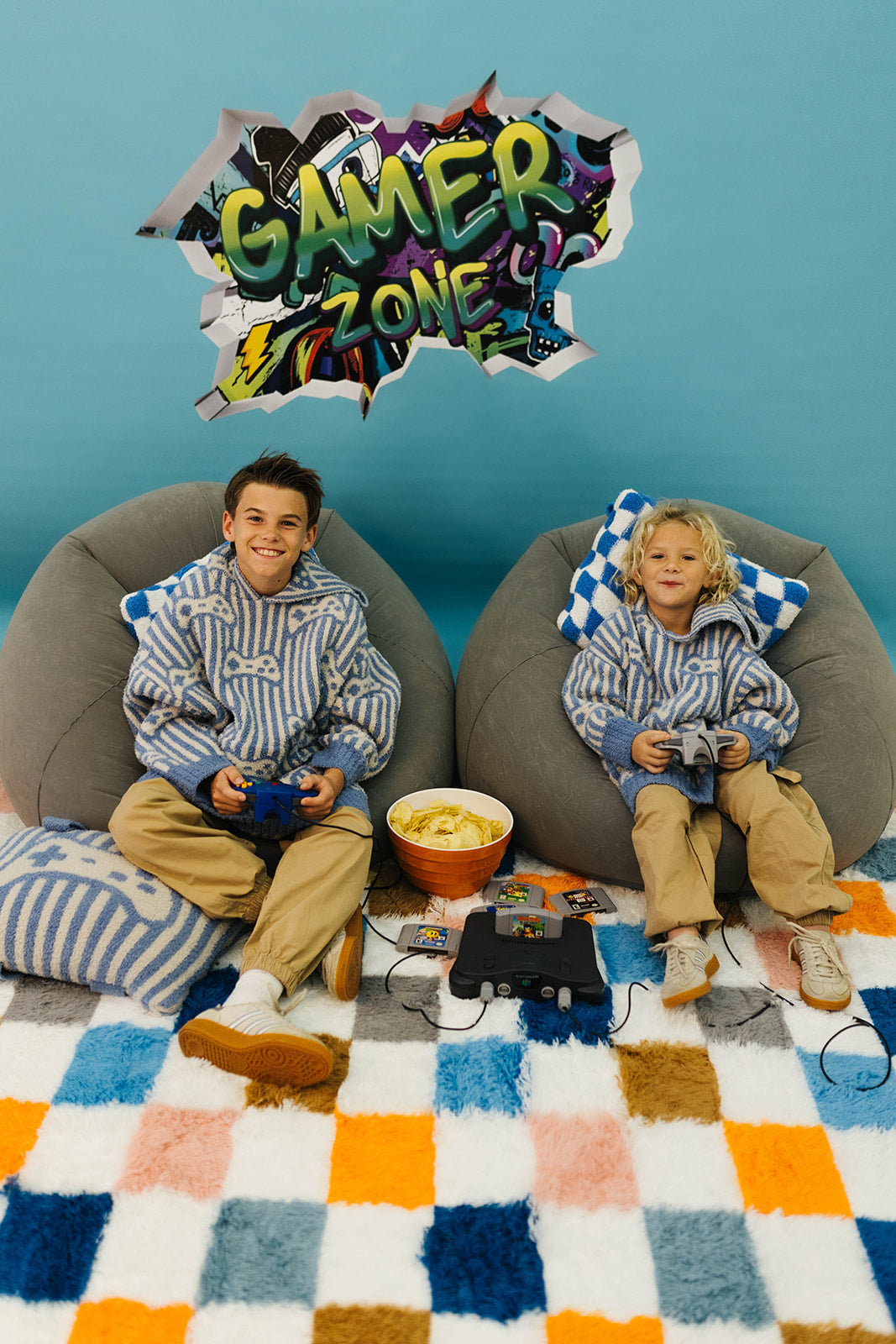 Two children sitting on bean bags with gaming equipment, surrounded by a colorful 'Gamer Zone' sign.
