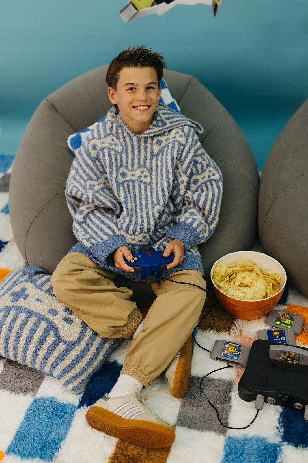 Child sitting on a bean bag chair with gaming console and snacks, wearing a blue and white striped sweater.