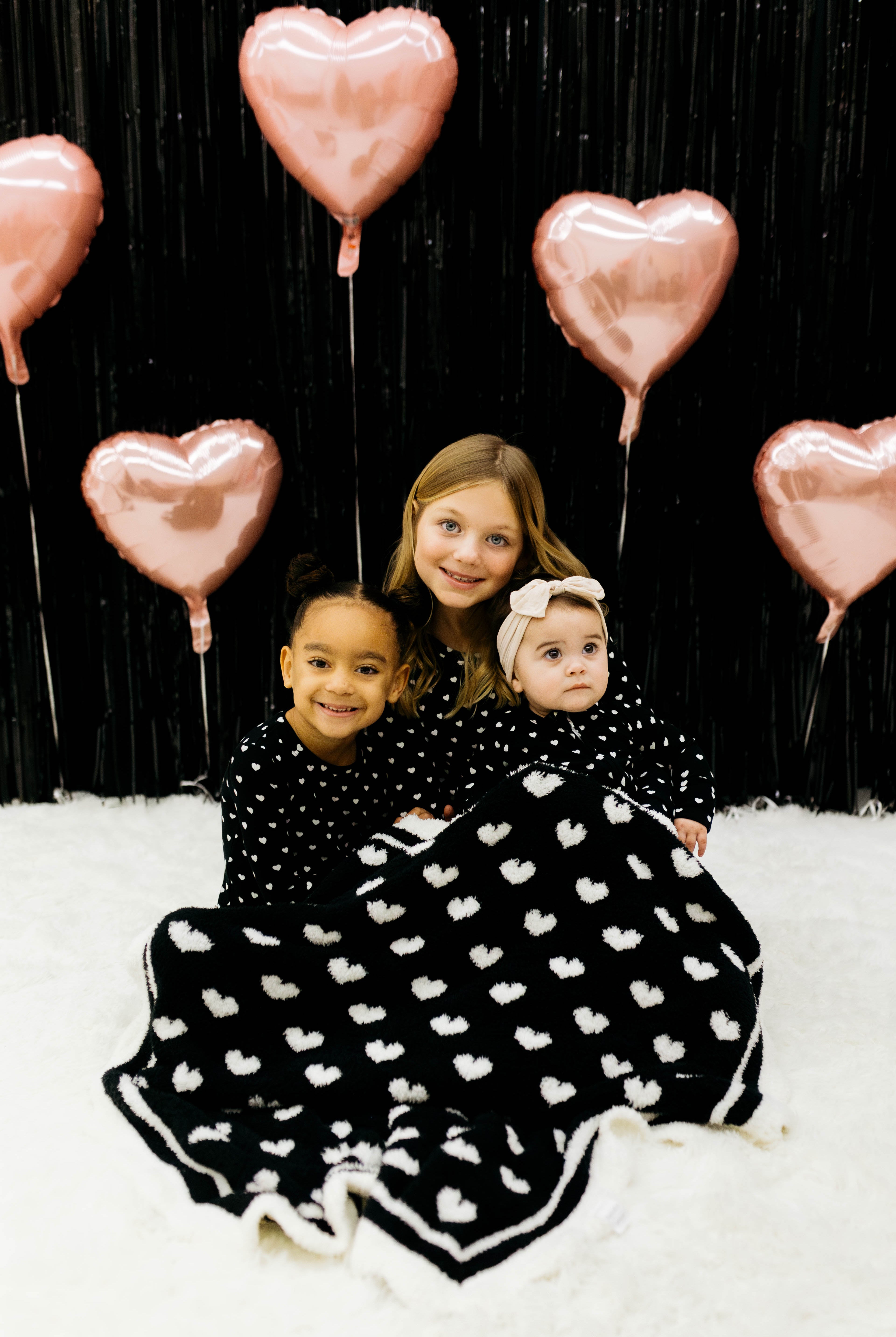 Three children in matching black and white pjs sitting on a white surface with pink heart balloons in the background. With the black blanket draped on them.