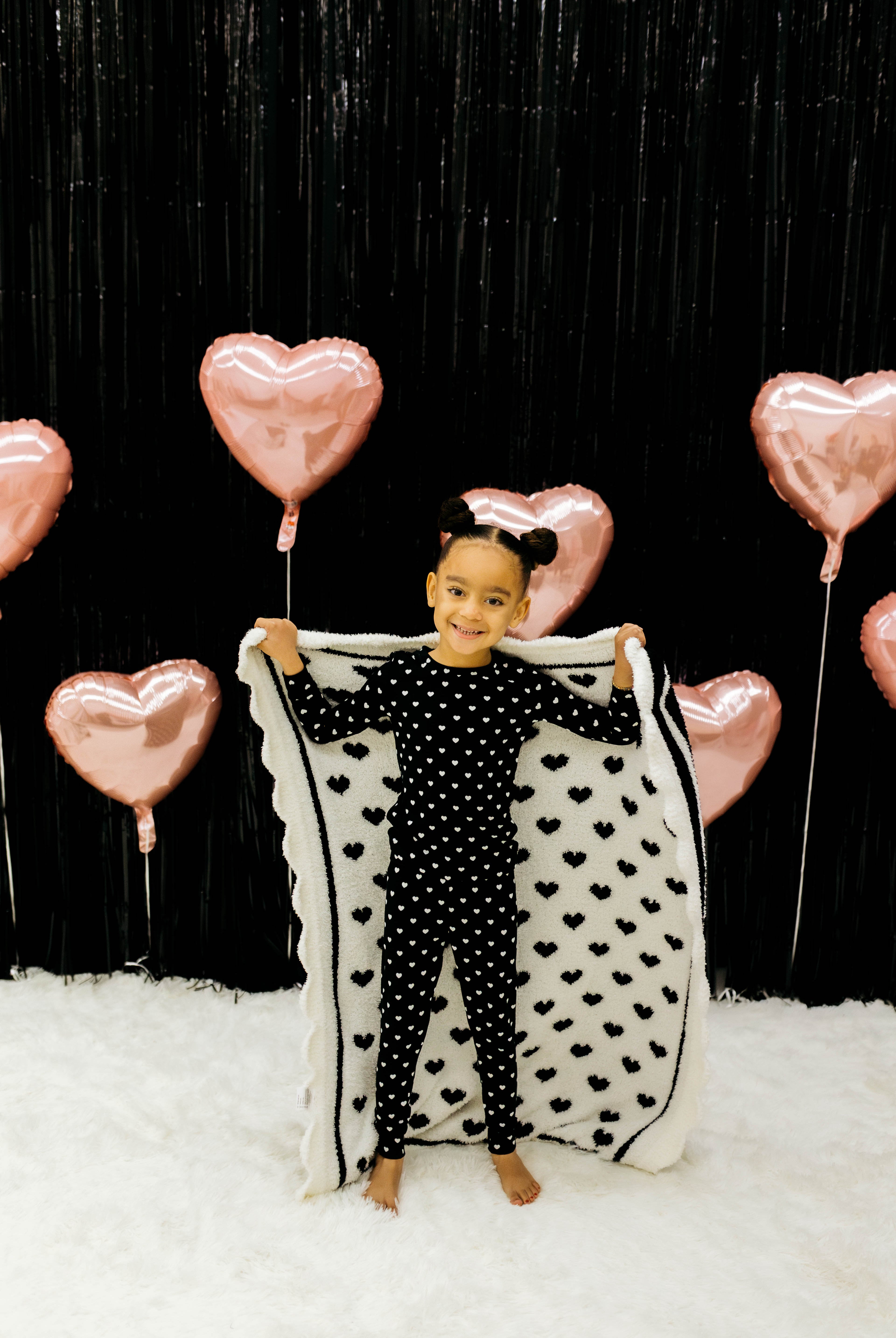 Child holding a heart-shaped blanket with pink heart balloons in the background