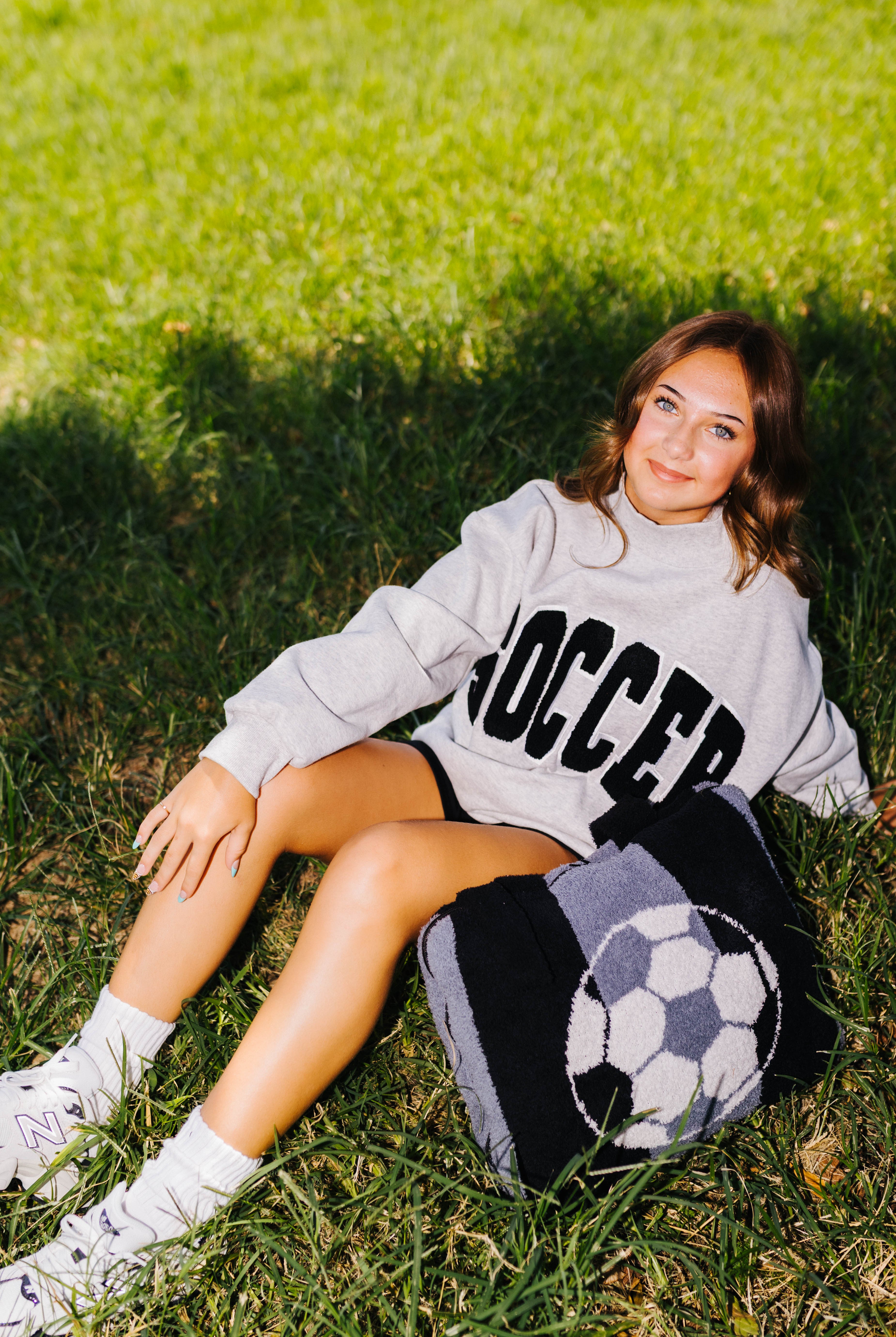 A young woman sits on a grassy lawn wearing a light gray sweatshirt with 'SOCCER' printed in black, black shorts, and white sneakers. A black and white soccer-themed knit quillow rests beside her.