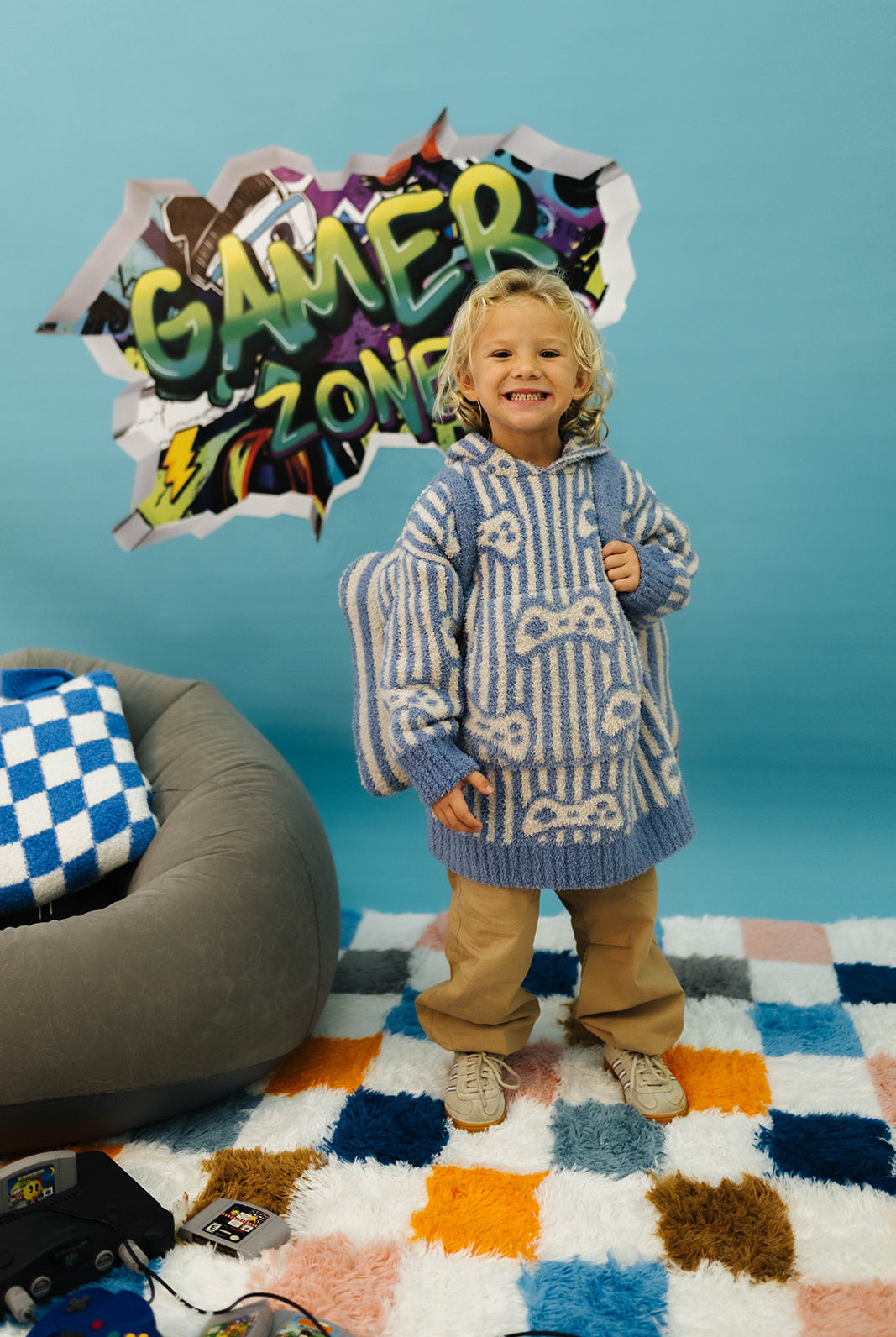 Child in a patterned sweater standing in front of a 'Gamer Zone' sign with a colorful rug and bean bag chair.