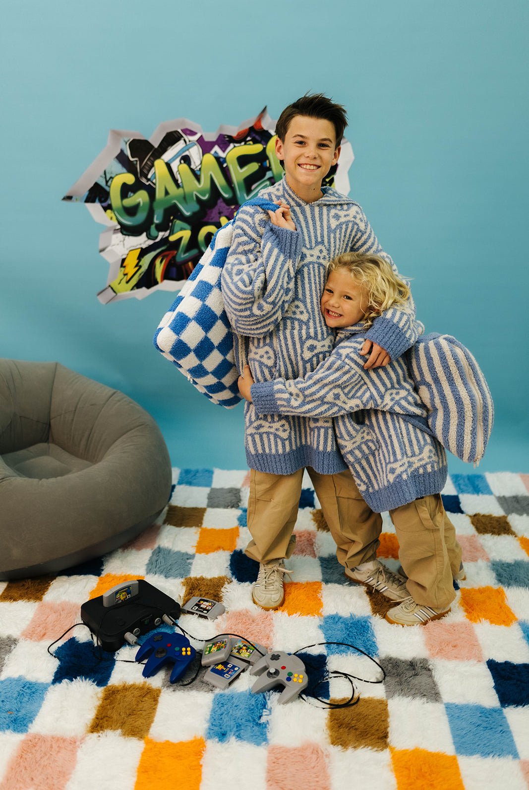 Two children in matching striped outfits standing on a colorful checkered floor with gaming equipment around.