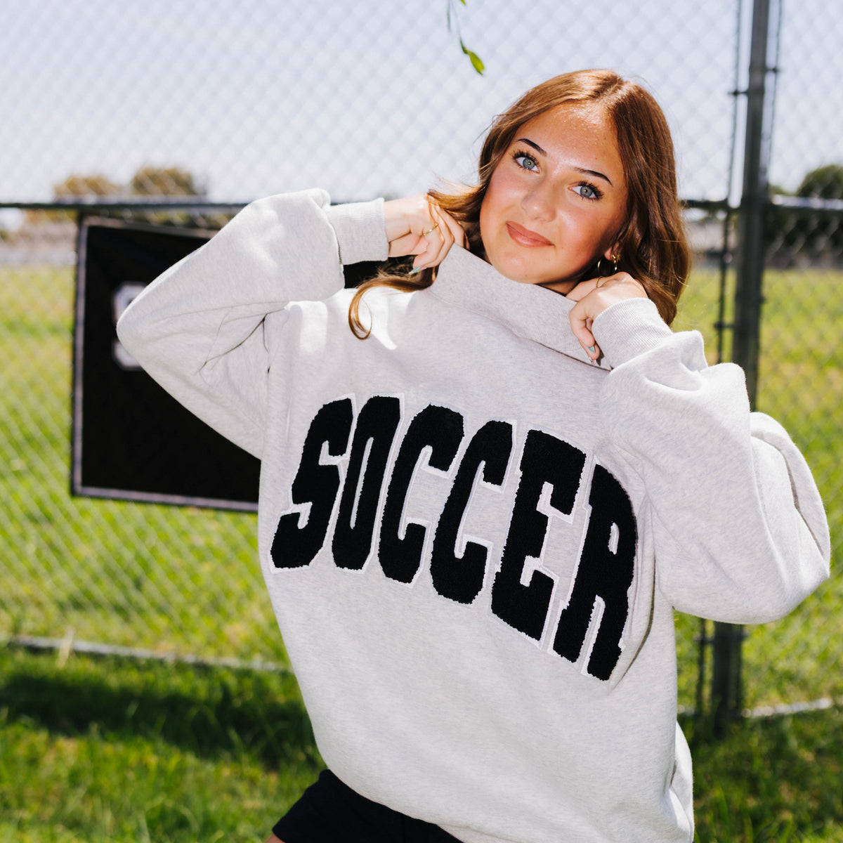 A woman posing outdoors in a gray crewneck sweatshirt with 'SOCCER' printed in black across the chest, paired with black athletic shorts, in front of a chain-link fence and grassy field.