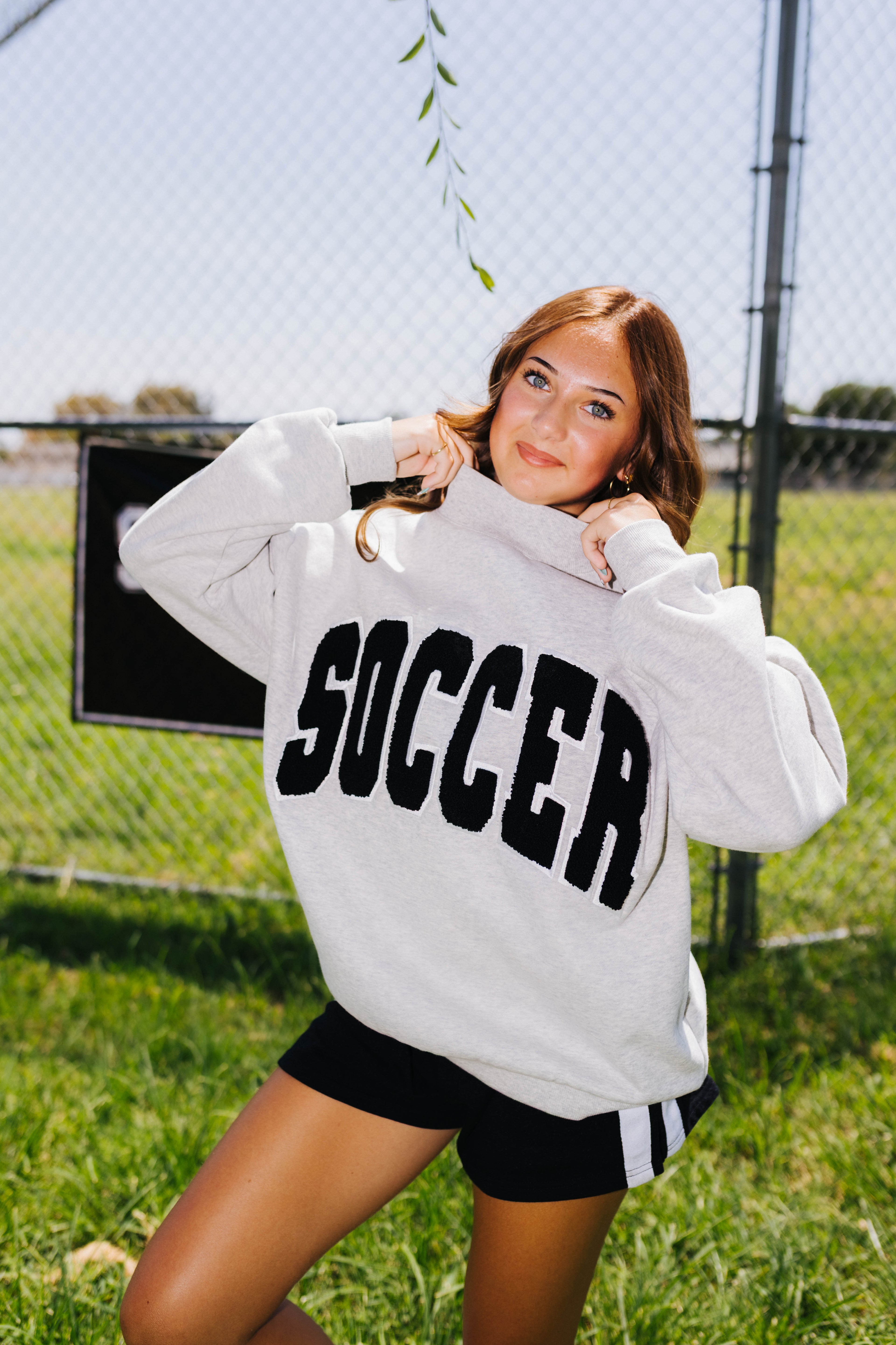 A woman posing outdoors in a gray crewneck sweatshirt with 'SOCCER' printed in black across the chest, paired with black athletic shorts, in front of a chain-link fence and grassy field.