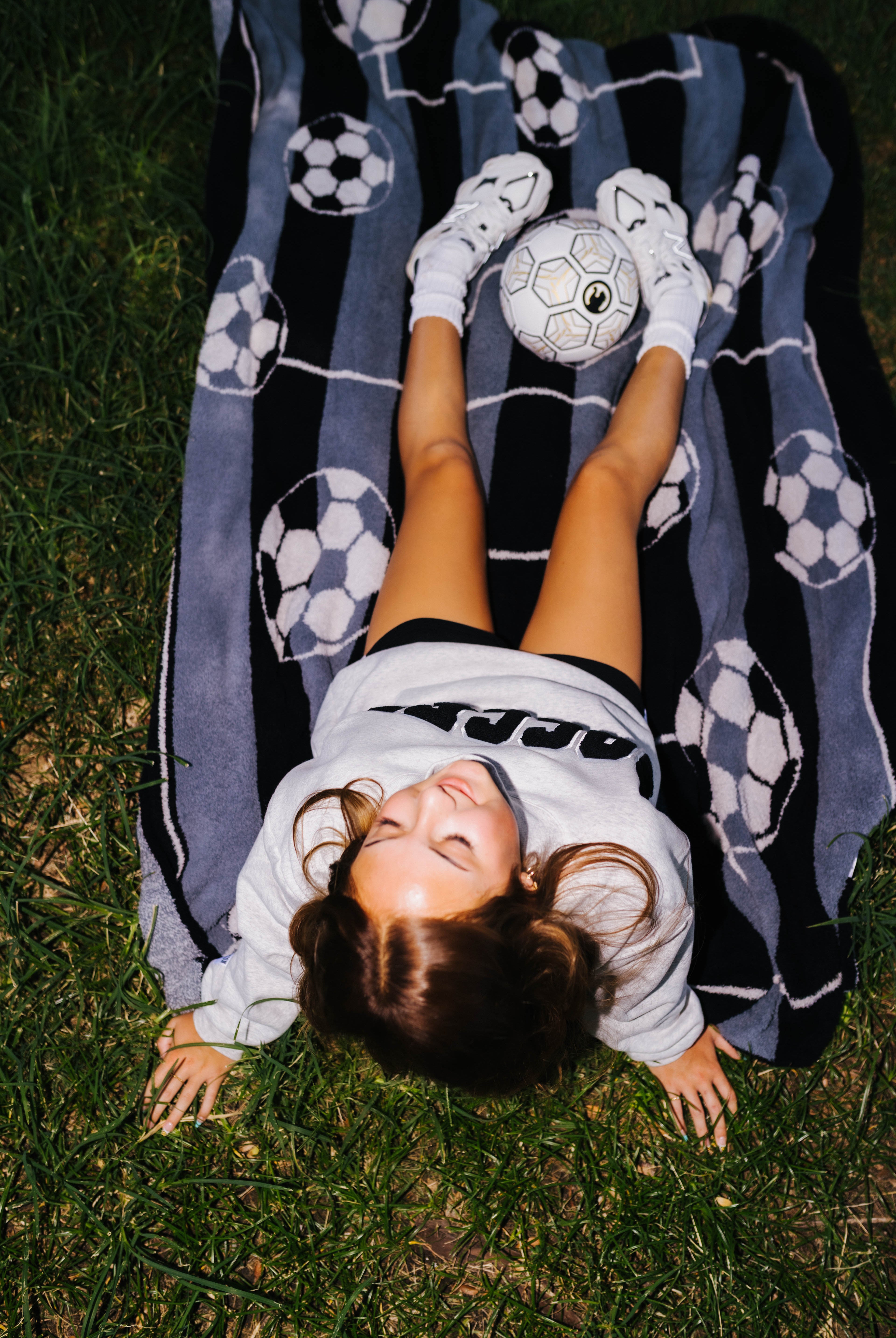 A girl lies on her back on green grass, wearing a gray sweatshirt, black shorts, and white socks, with a soccer ball on her feet. She is lying on a black and gray striped blanket with soccer ball patterns.