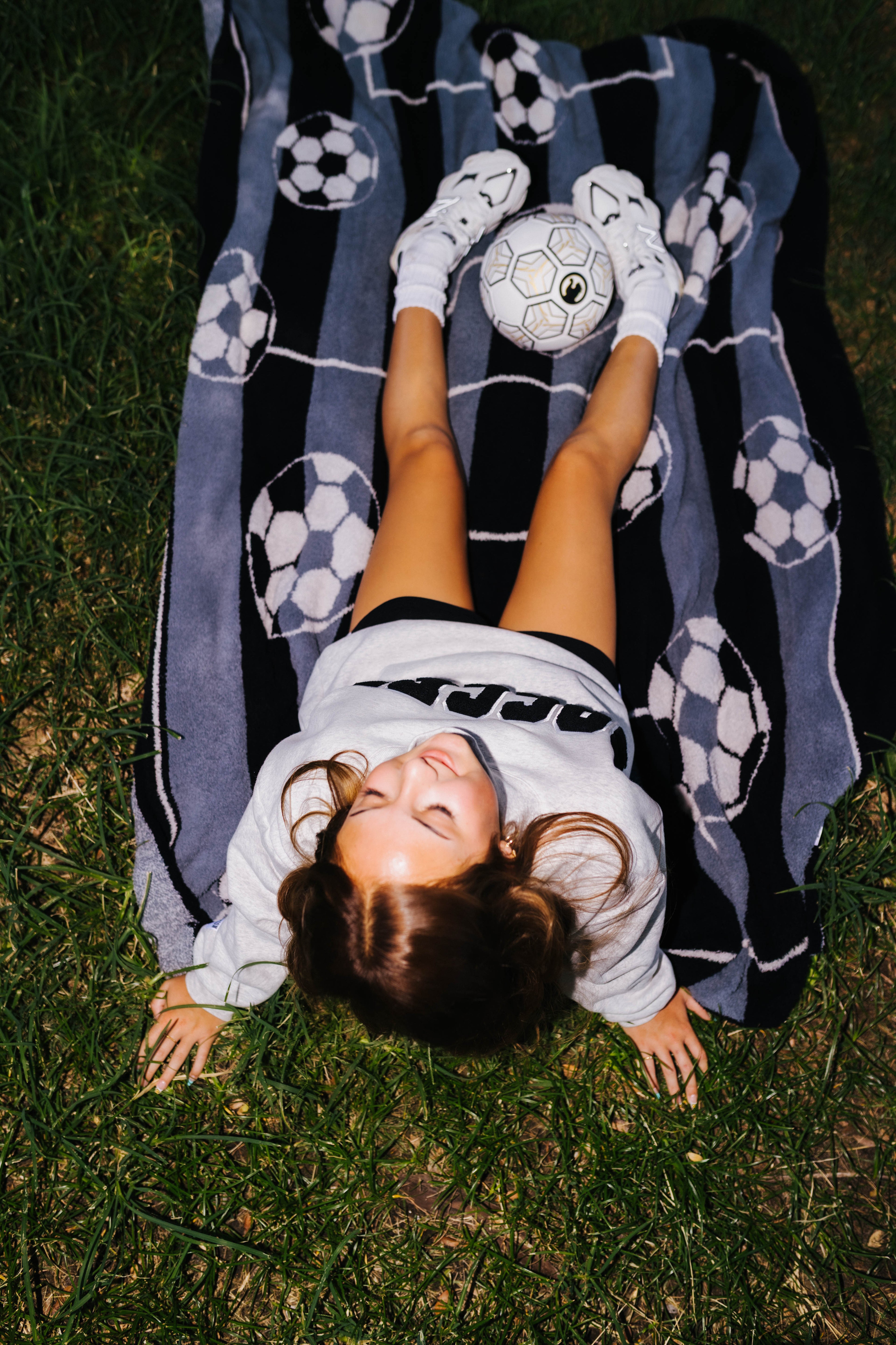 A girl lies on her back on green grass, wearing a gray sweatshirt, black shorts, and white socks, with a soccer ball on her feet. She is lying on a black and gray striped blanket with soccer ball patterns.