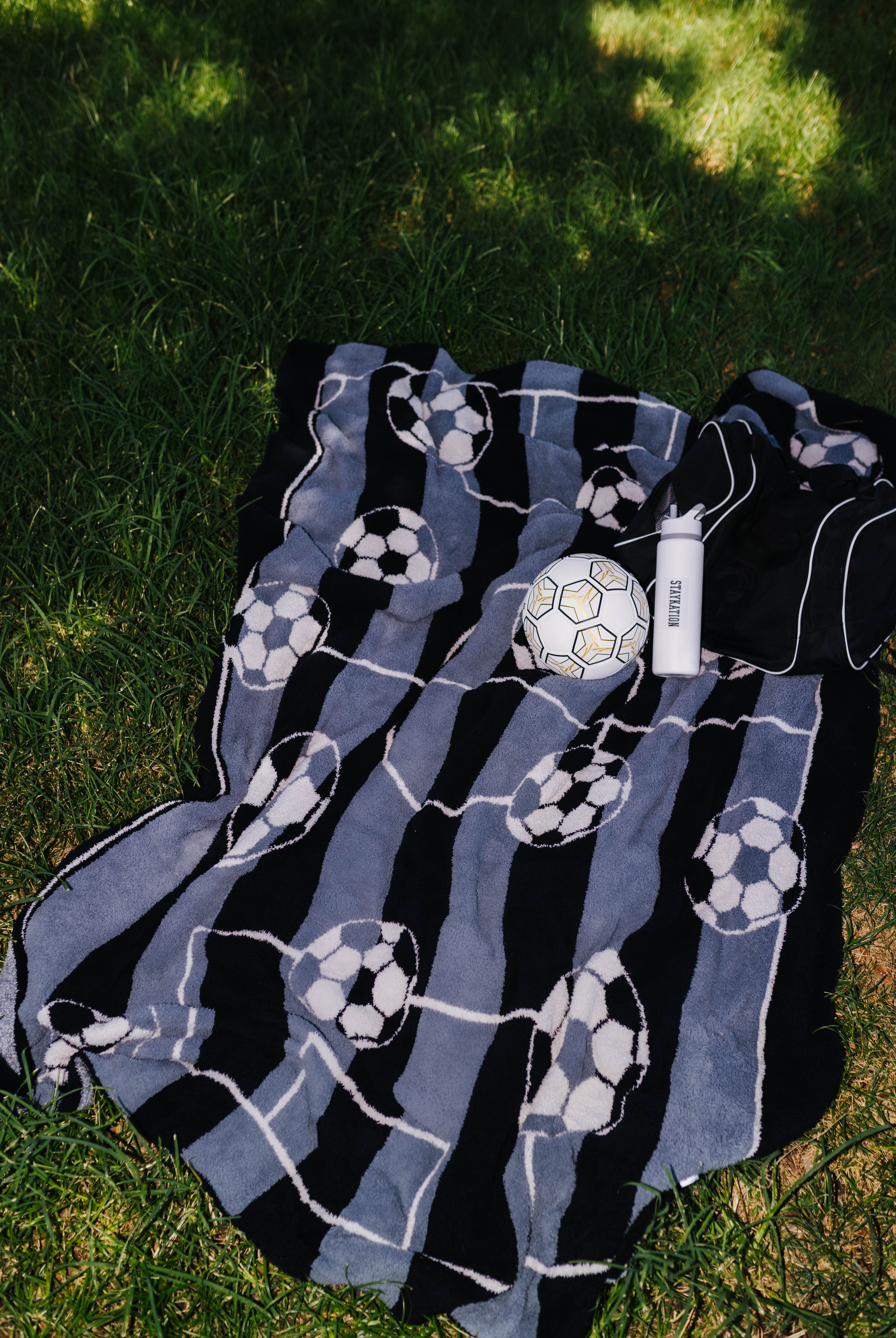 A black and blue striped blanket with white soccer ball patterns, laid out on green grass next to a white soccer ball.