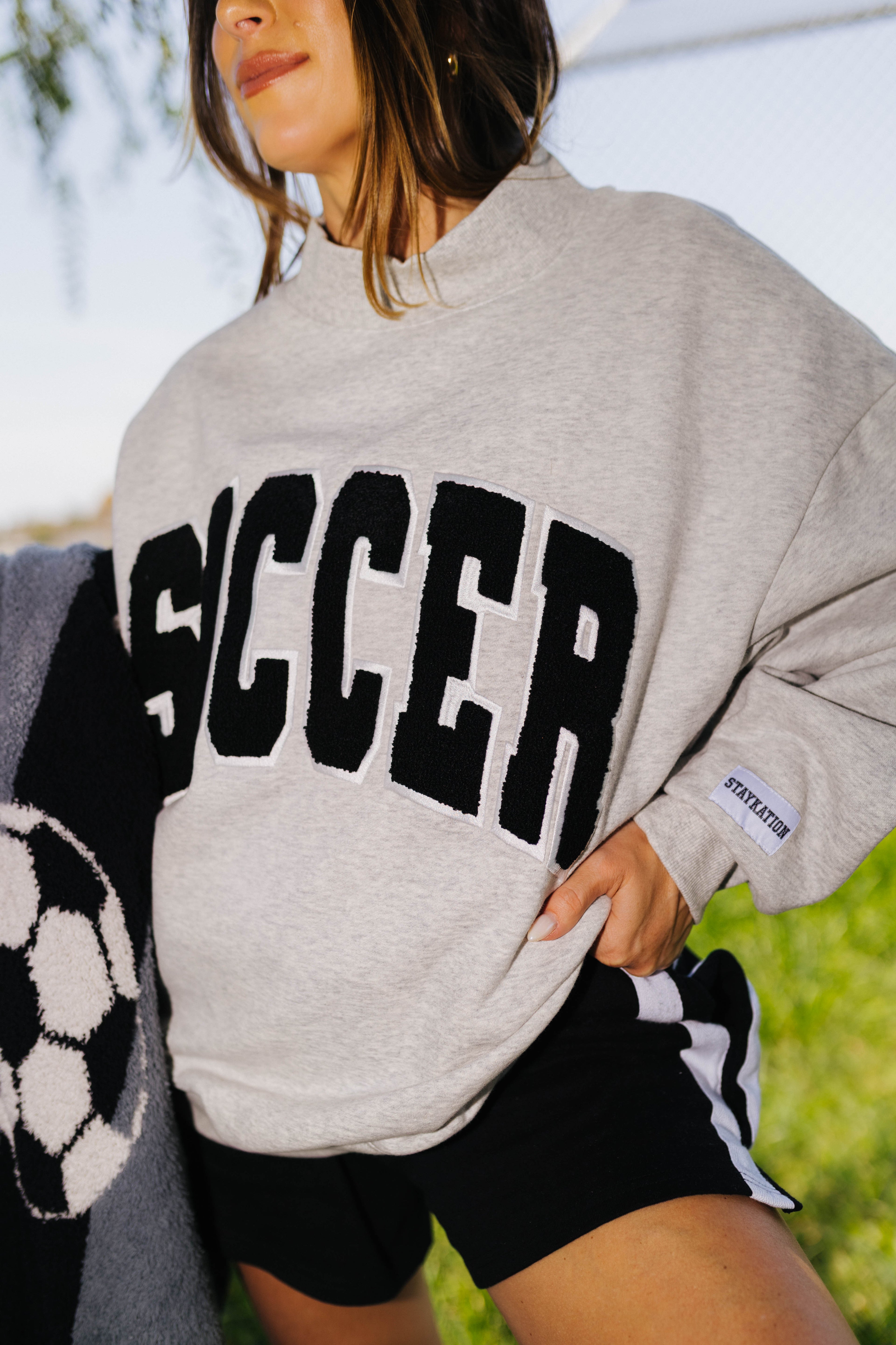 A woman wearing an oversized light gray knit sweatshirt with the word 'SOCCER' in large black varsity-style letters across the front, paired with black athletic shorts with white side stripes. The photo is taken outdoors with a blurred green and blue background.