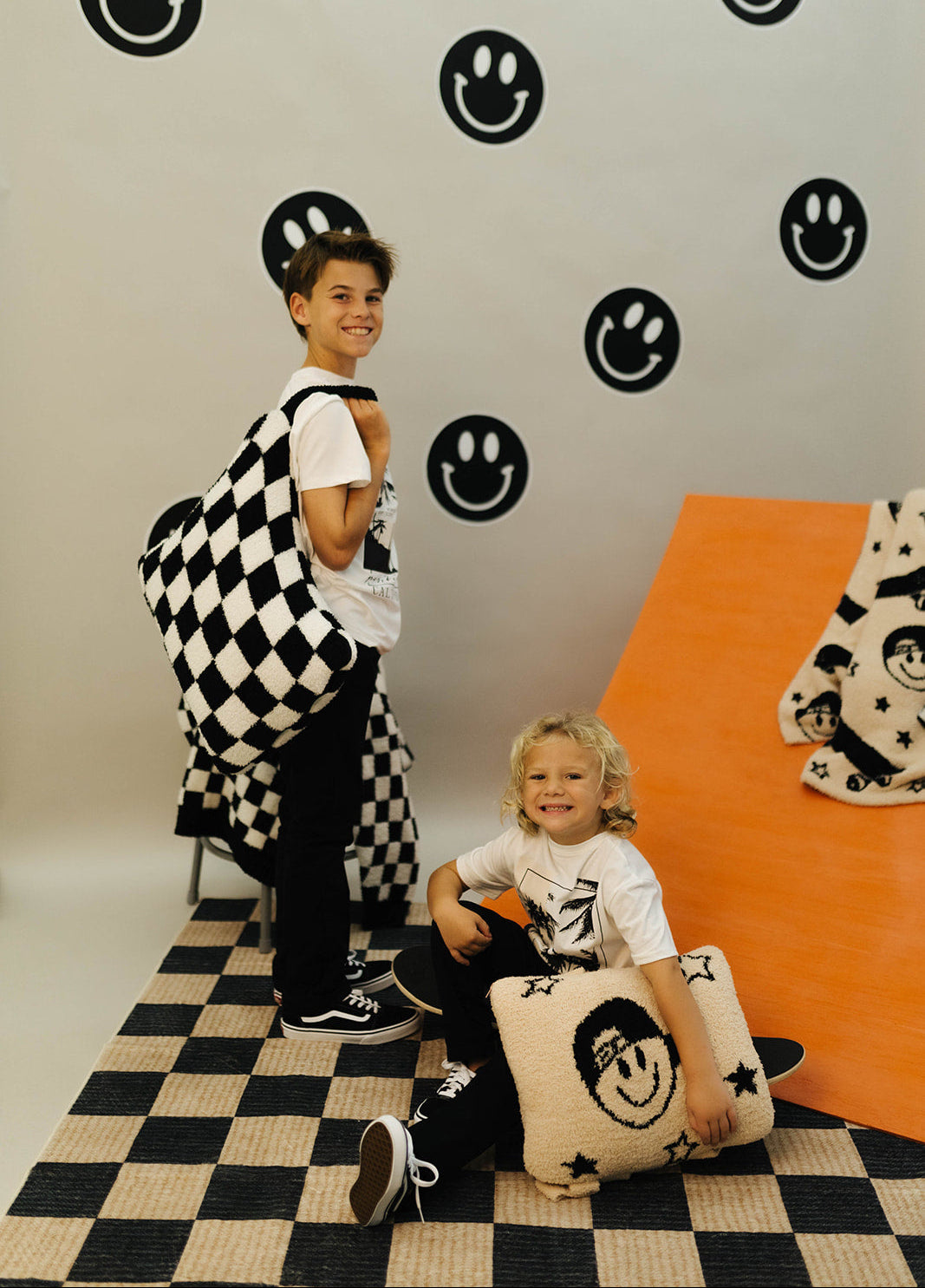 Two children in a room with checkered floor and wall decorations featuring smiley faces.