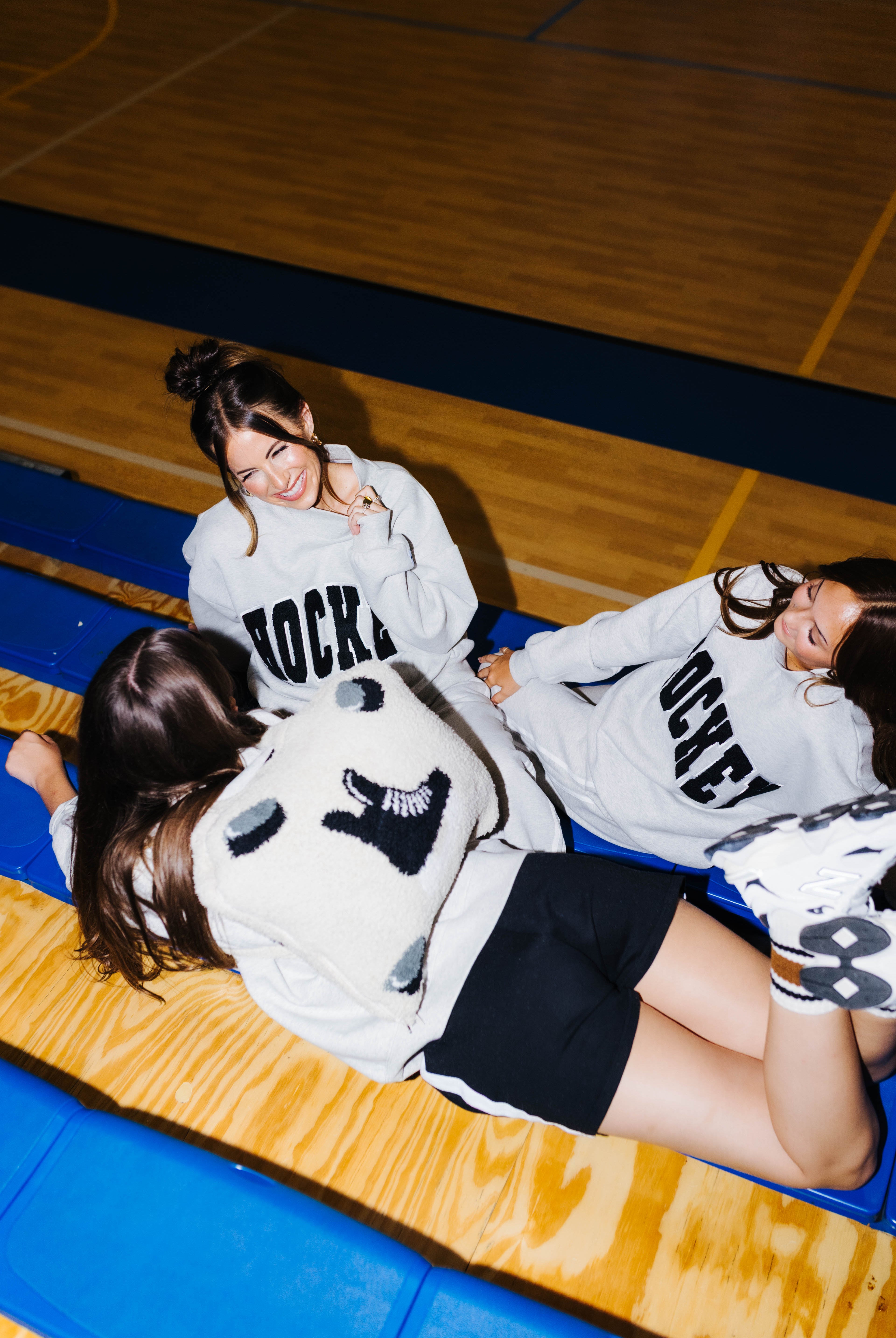 Three women lying on blue gym bleachers in a basketball court, wearing matching graphic sweatshirts with 'Hockey' printed in black.