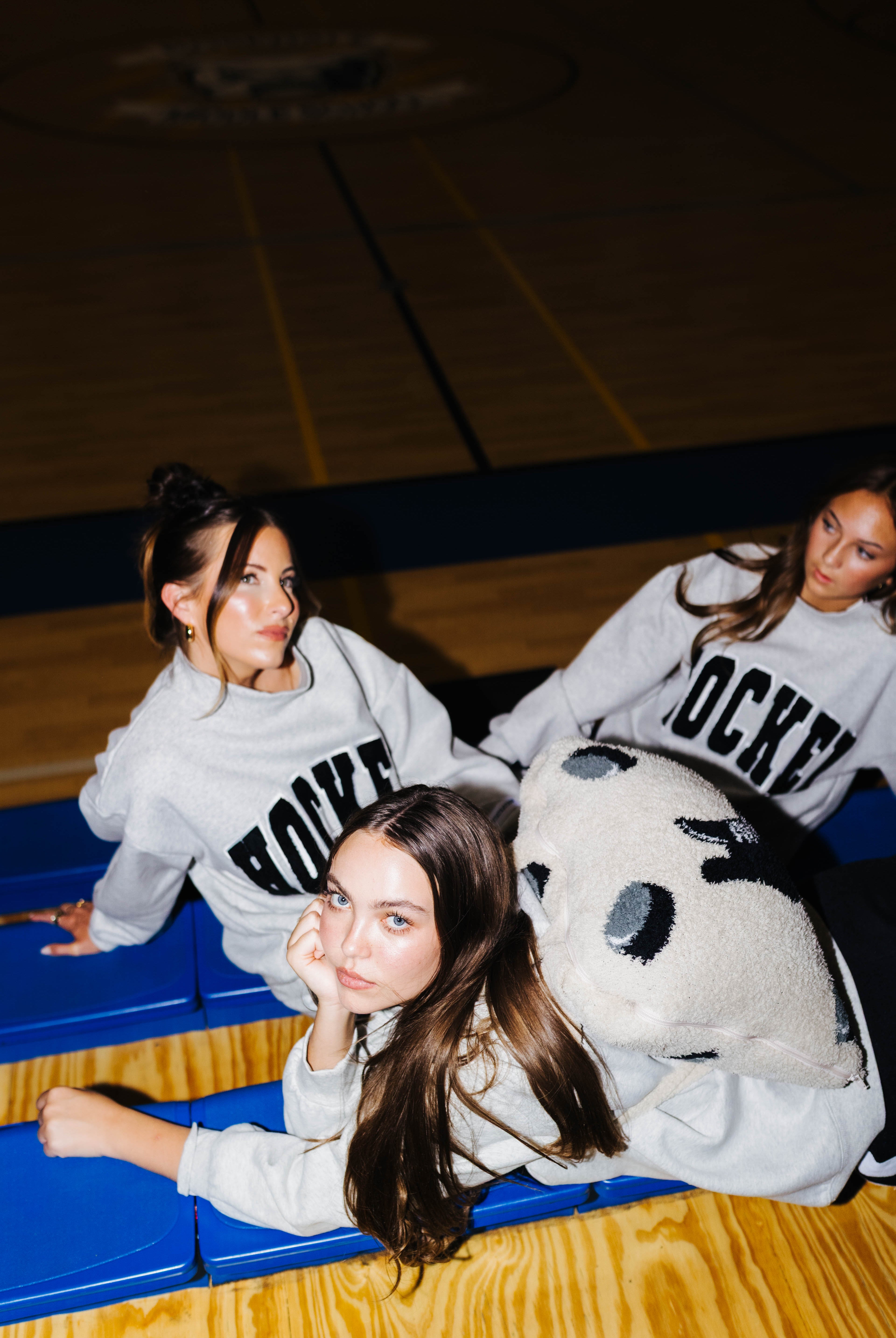 Three women in matching light gray crewneck sweatshirts with 'HOCKEY' printed in black across the chest, posed on blue gym bleachers in a basketball court.