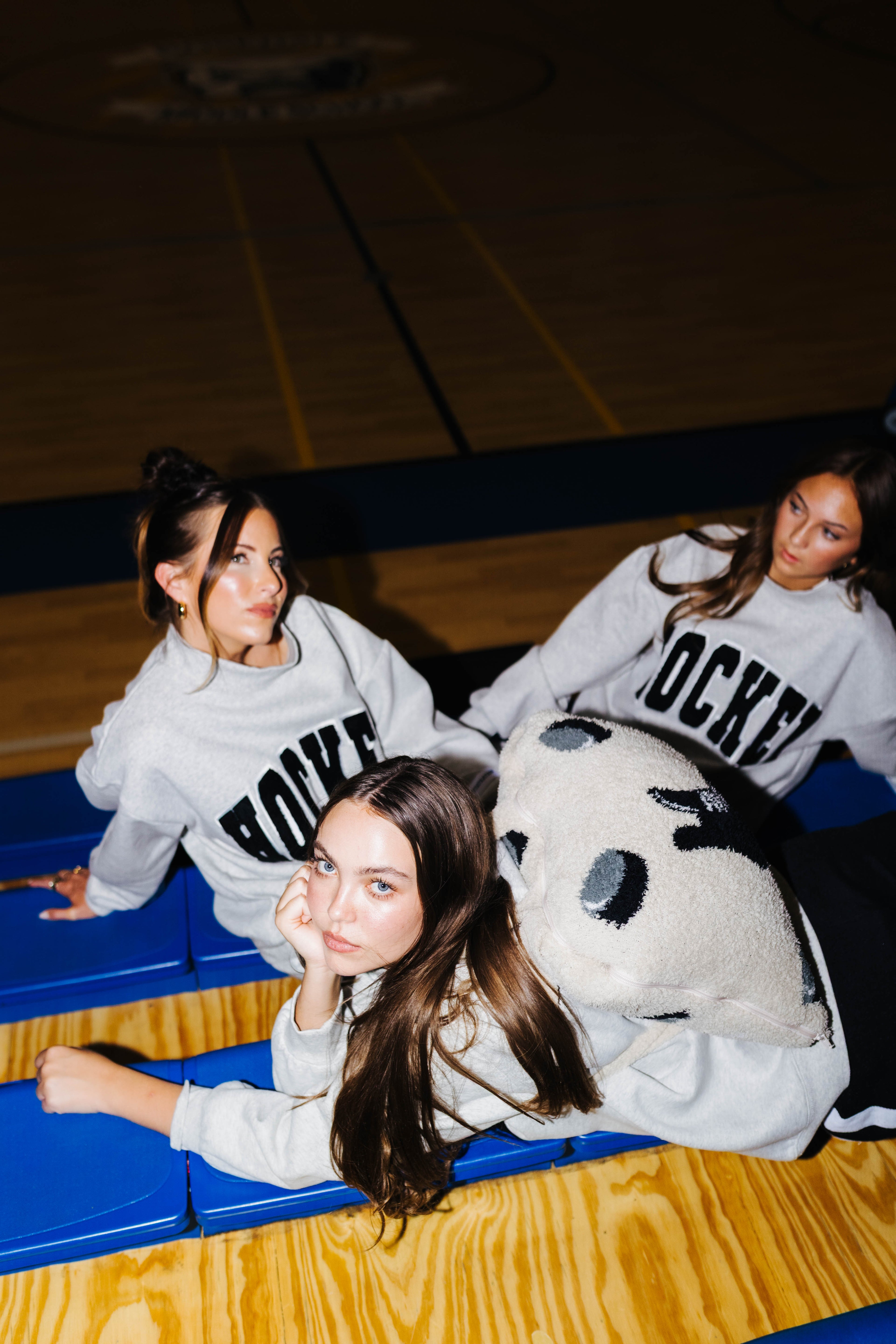 Three women in matching light gray crewneck sweatshirts with 'HOCKEY' printed in black across the chest, posed on blue gym bleachers in a basketball court.