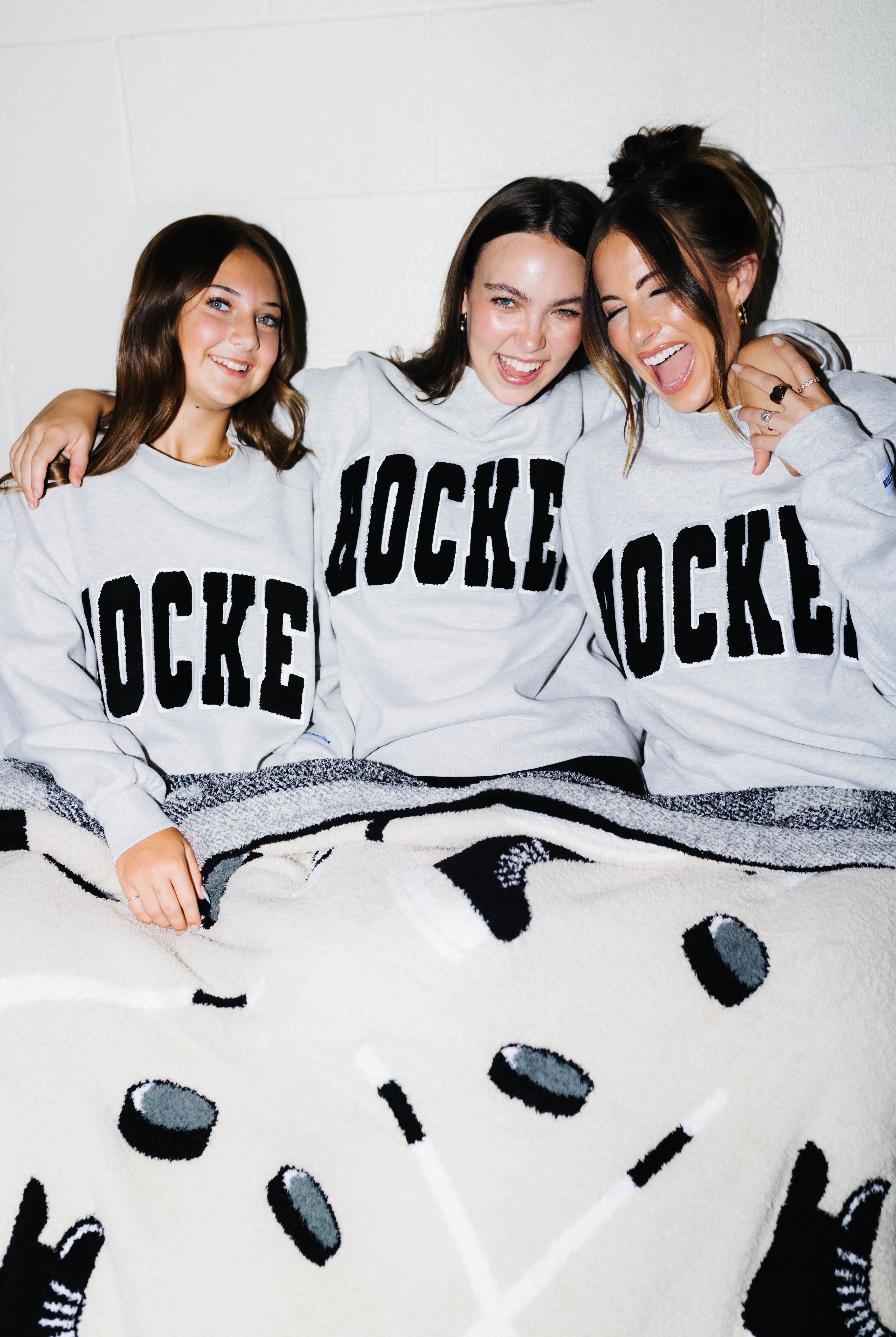 Three women sitting together on bleachers wearing matching light gray hoodies with 'HOCKEY' printed in black varsity-style letters across the front. A cream knit blanket with black hockey motifs is spread across the bed.