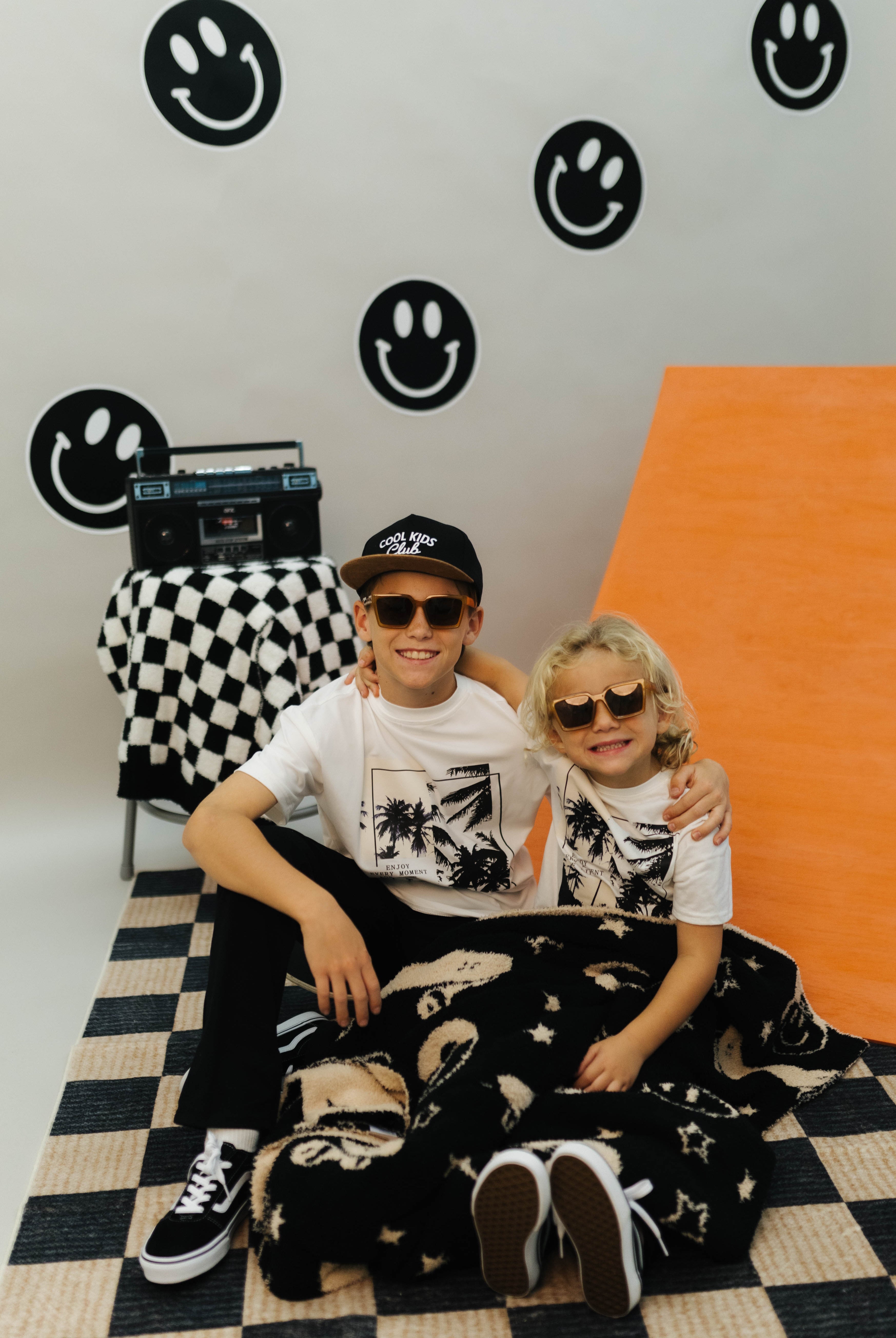 Two children sitting on a checkered floor with a wall featuring smiley face designs.
