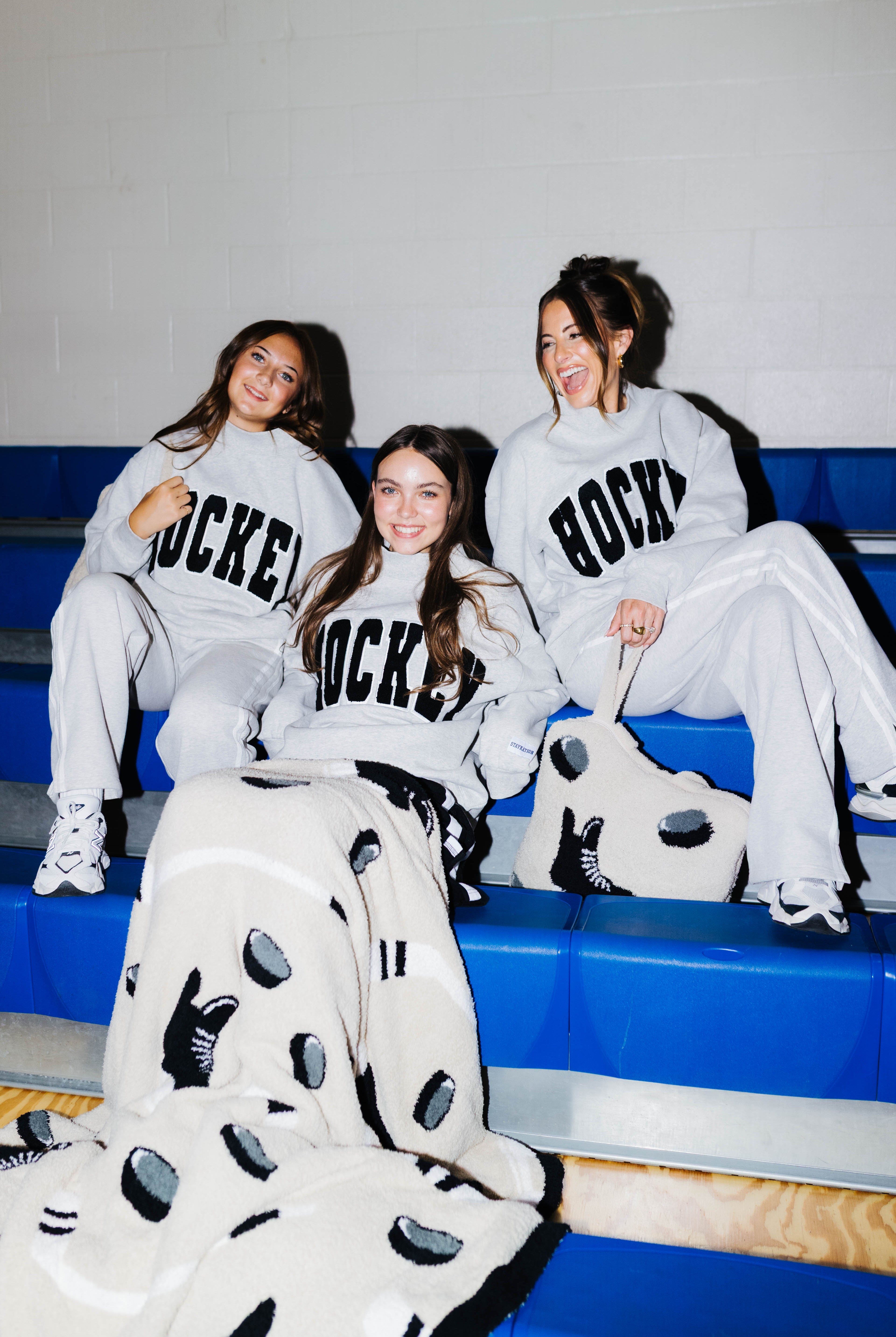Three women sitting on blue gym bleachers wearing matching gray hockey sweatpants with black HOCKEY lettering with the blanket draped over them